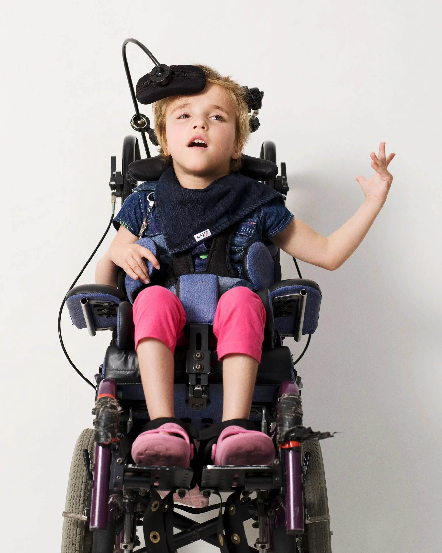 Portrait of a girl (Rachel) wiht cerebral palsy sitting in a wheel chair with pink trousers and pink shoes gesturing with her left hand by photographer David.A.Knight ©