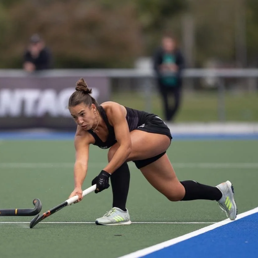 Female field hockey player crouched on the field, reaching to hit the ball with her stick during a game, wearing athletic gear and gloves.
