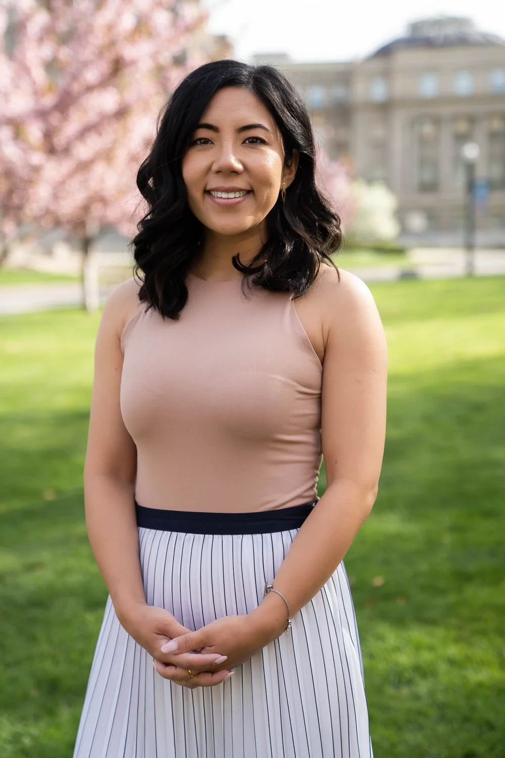 A young woman with dark wavy hair smiling outdoors in a park with pink cherry blossoms and a historic building in the background, wearing a sleeveless beige top and a striped skirt.