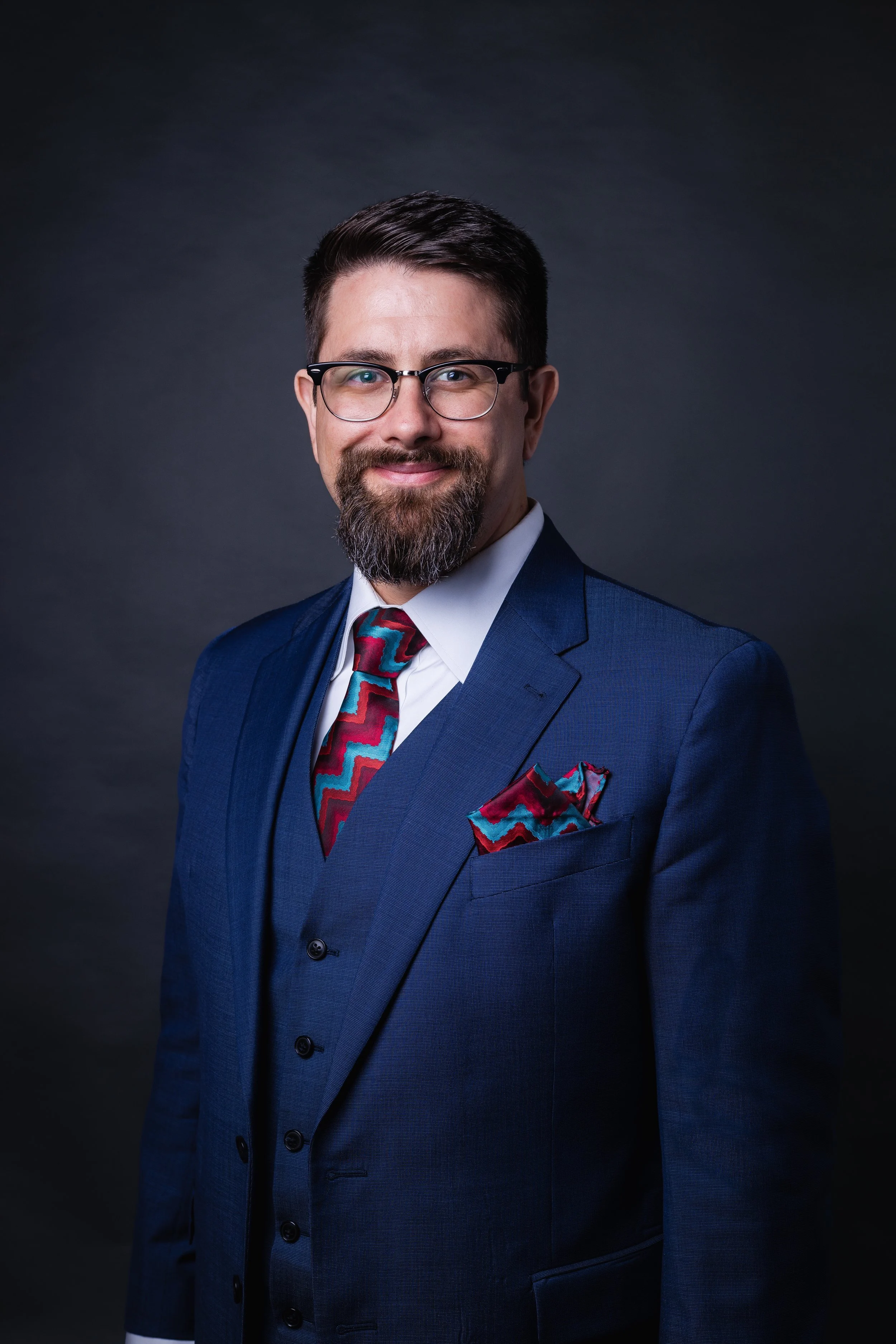 A man in a dark blue suit, white shirt, and colorful zigzag patterned tie and pocket square, wearing glasses, smiling against a dark background.