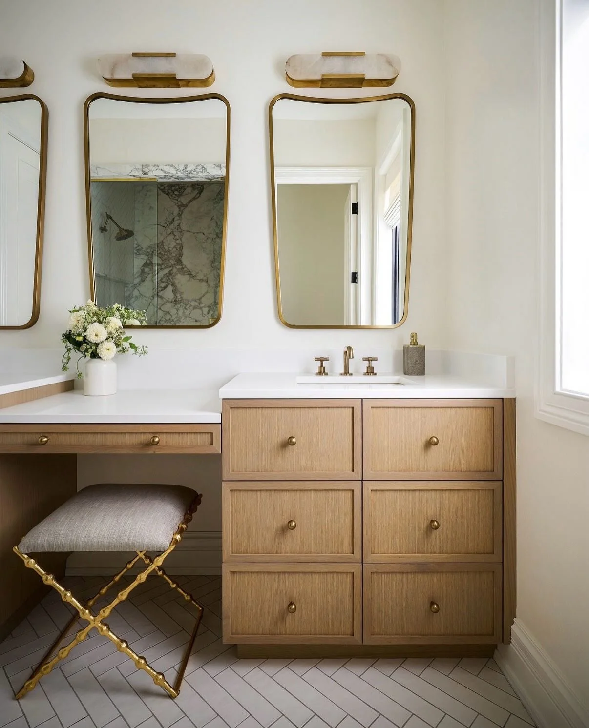 Subtle in its palette yet rich in character, this Lincoln Park primary bathroom combines custom white oak cabinetry, dramatic Arabescato marble, herringbone tile floors, natural stone lighting, and refined gold accents. A trio of softly curved mirror