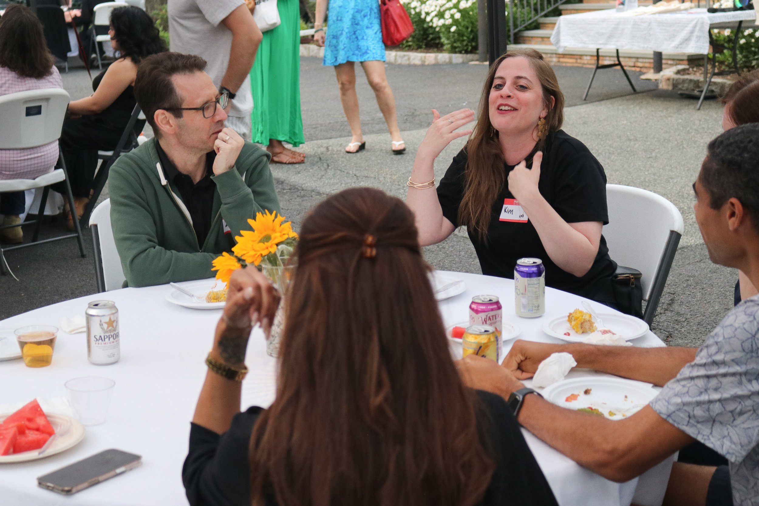 Group of people sitting around a white table outdoors at a social gathering, with some eating and drinking, decorated with a sunflower centerpiece, engaging in conversation.