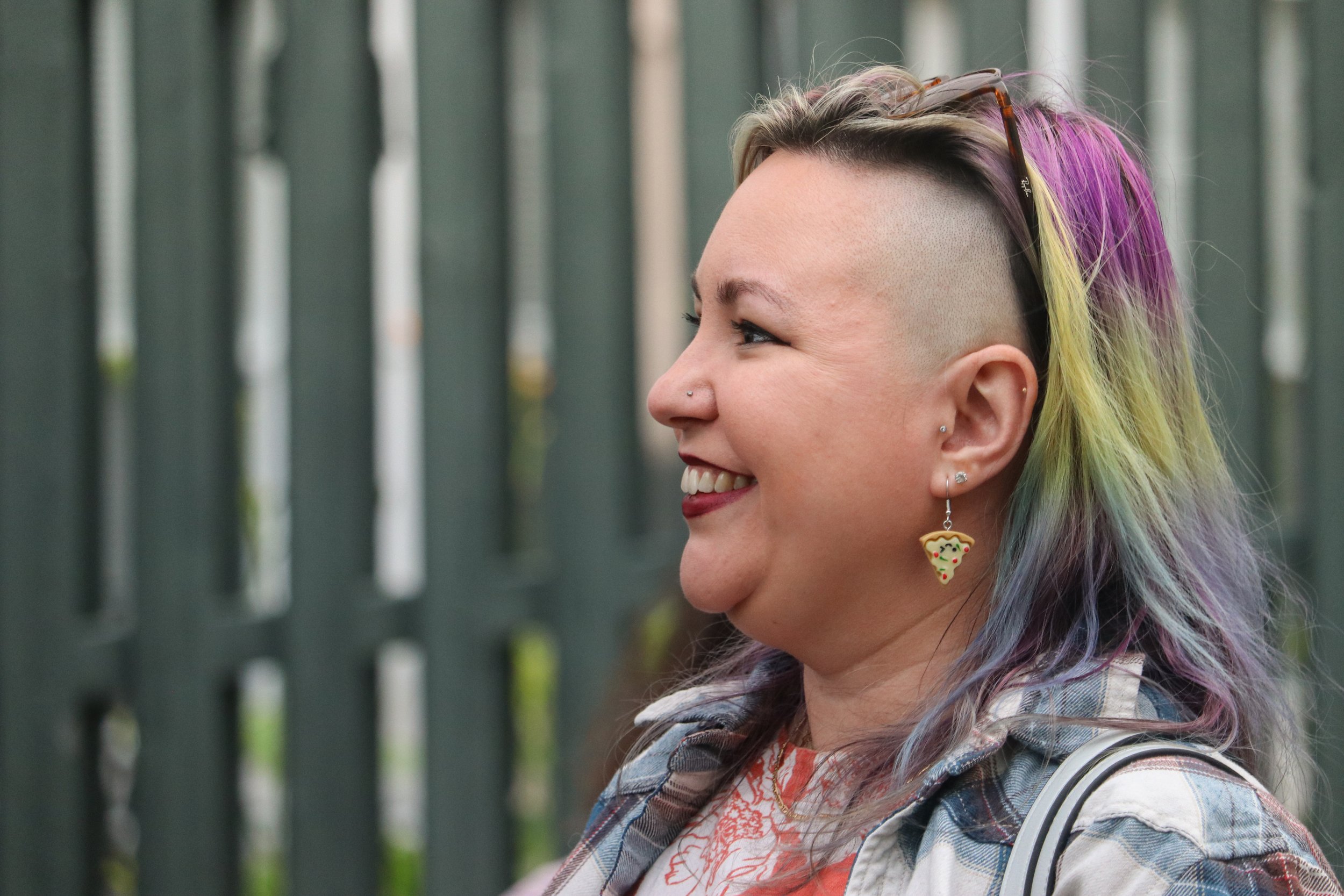 Side profile of a woman with colorful rainbow hair, wearing pizza slice earrings and smiling, outdoors near a green fence.