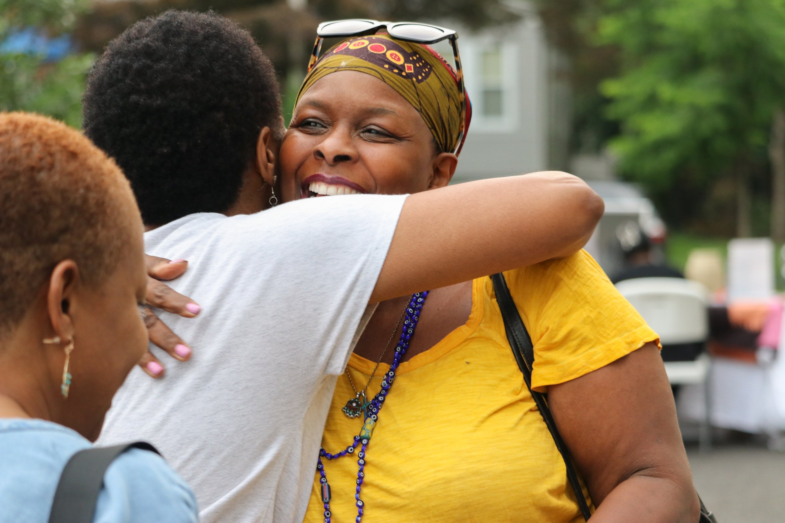 Three women hugging outdoors, smiling, with a background of trees and parked cars.
