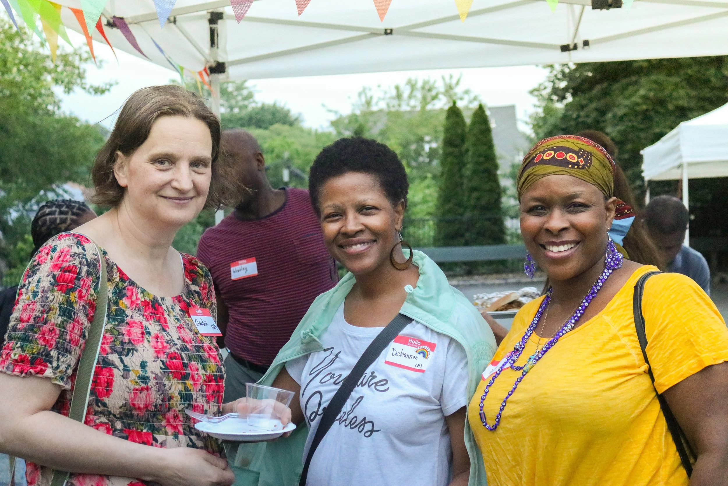 Three women at an outdoor event standing under a white canopy with colorful triangular bunting. They are smiling, wearing casual clothing, and have name tags on. One woman is holding a clear plastic cup.