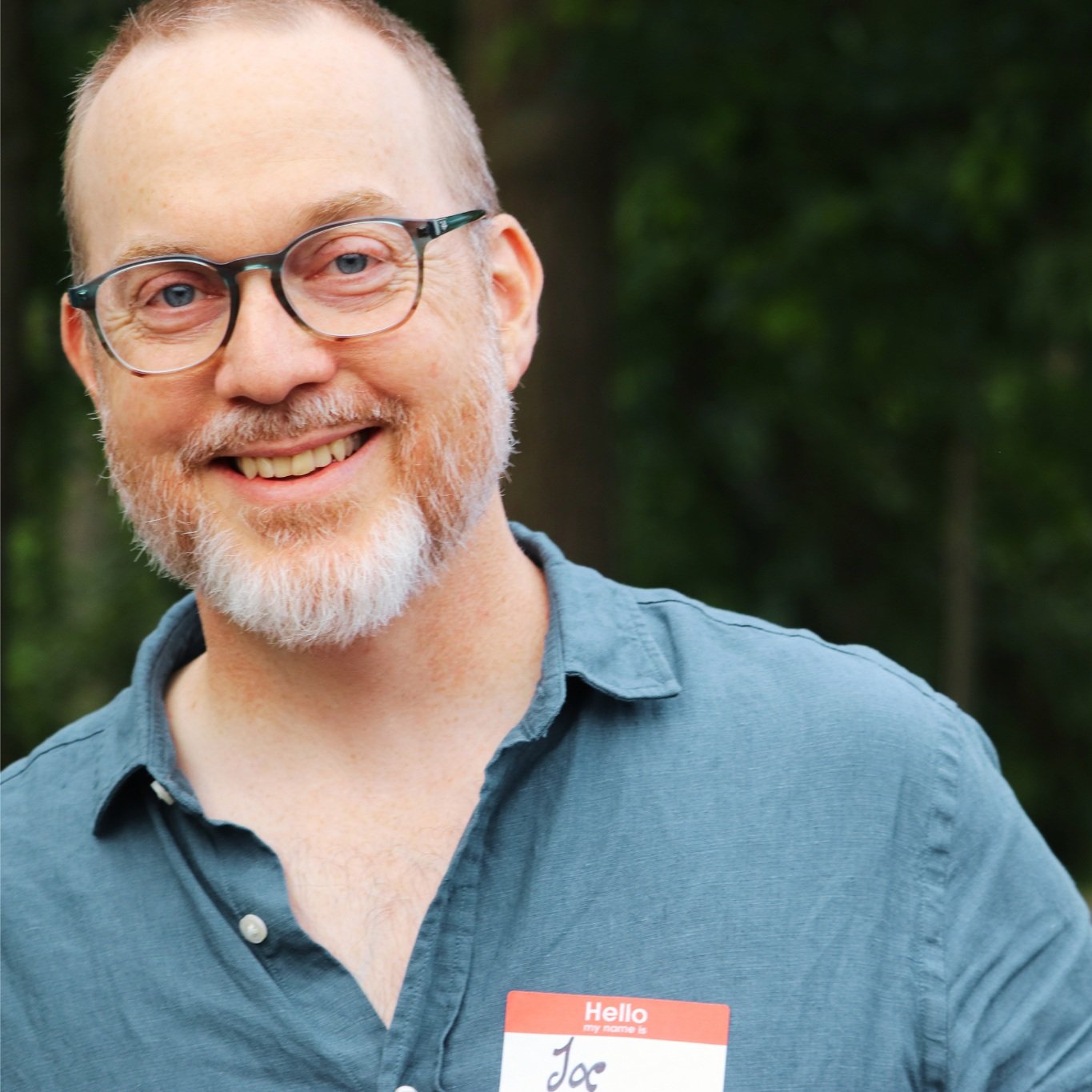 A smiling man with glasses, a beard, and a name tag that reads 'Hello my name is Jac,' standing outdoors with trees in the background.