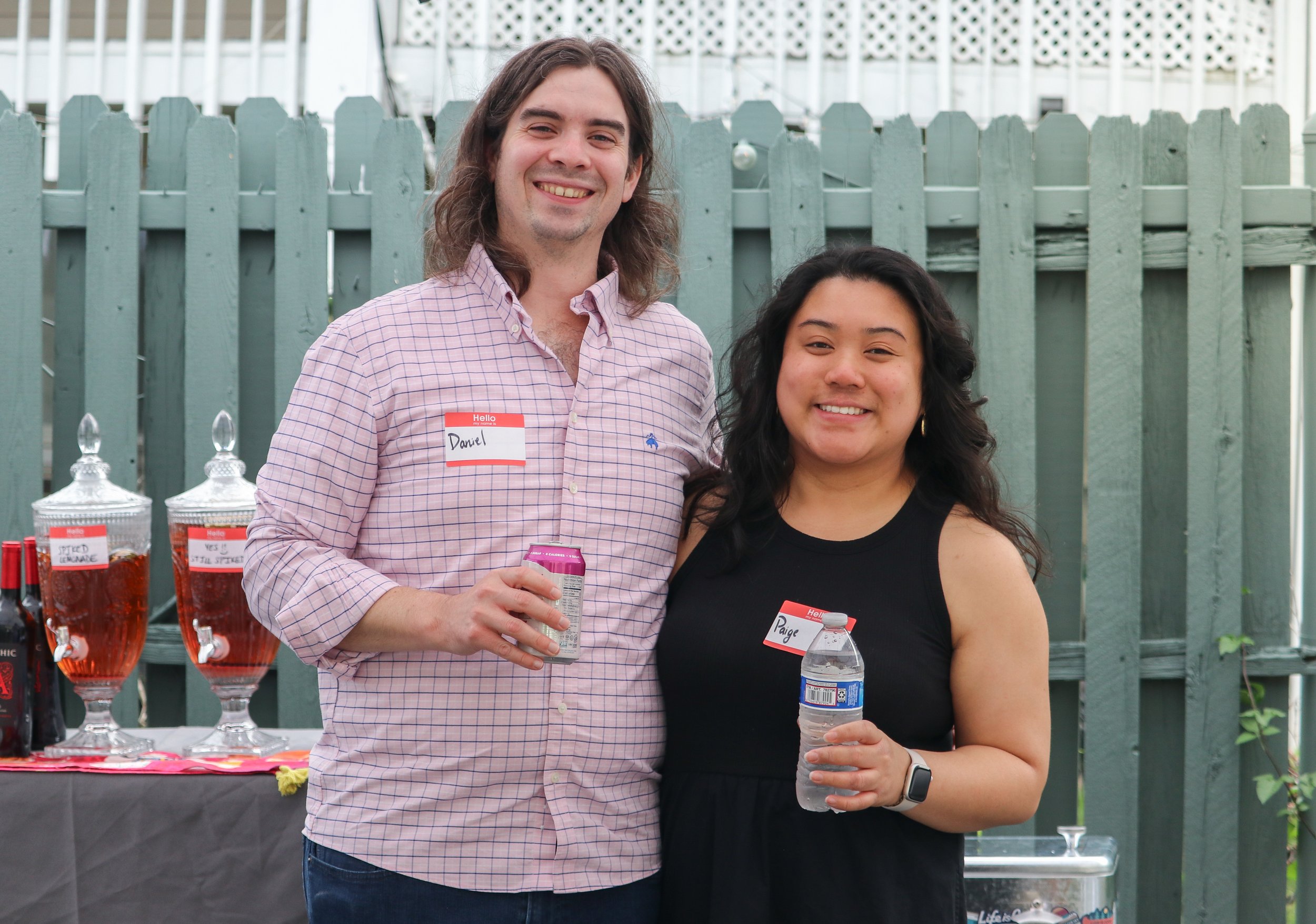 A man and a woman smiling and holding drinks at an outdoor event, standing in front of a wooden fence, with a table of beverages behind them. Both are wearing name tags, with the man’s name being Daniel and the woman’s Paige.