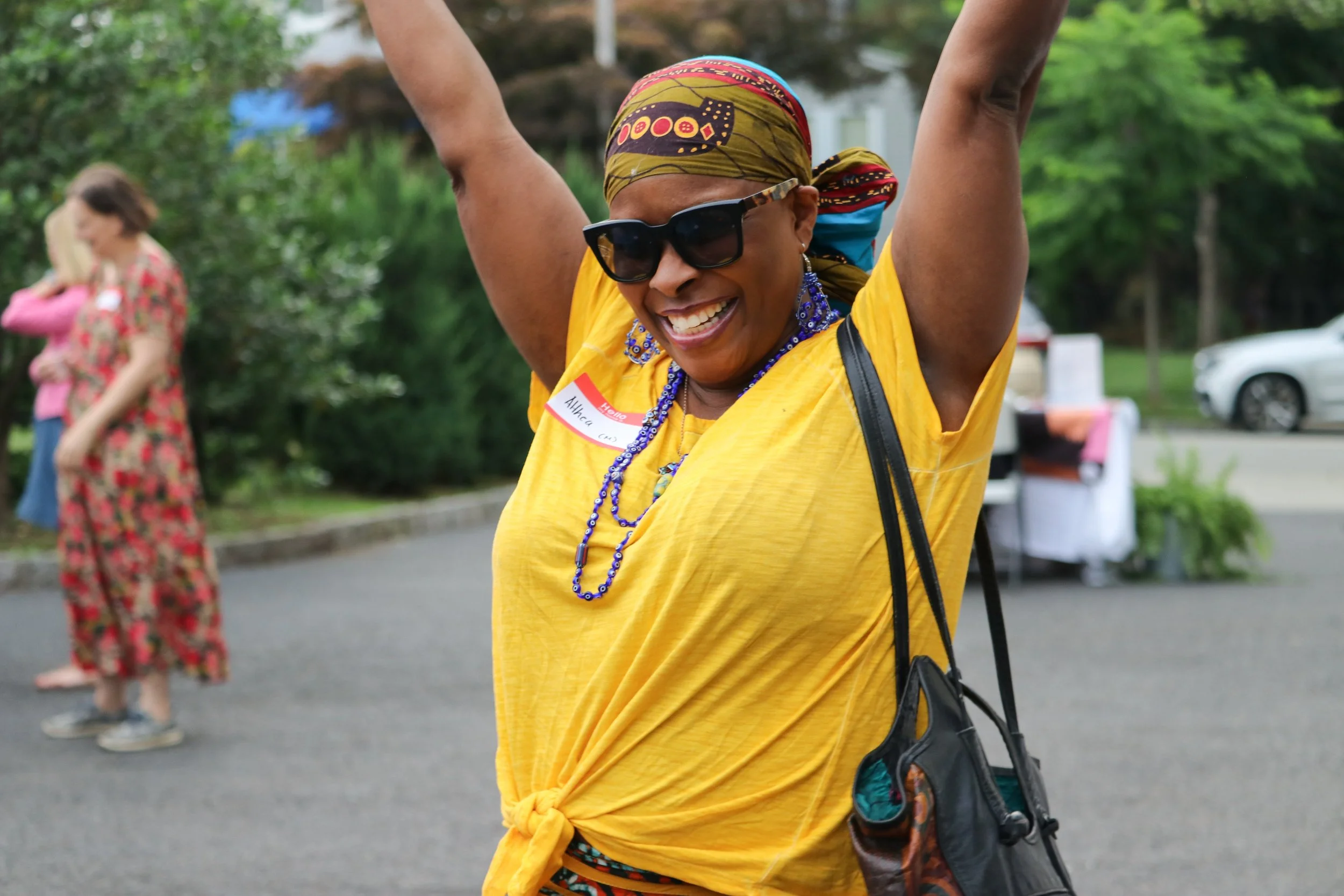 A smiling woman in a yellow shirt, sunglasses, and colorful headscarf, raising her arms in joy at an outdoor event.
