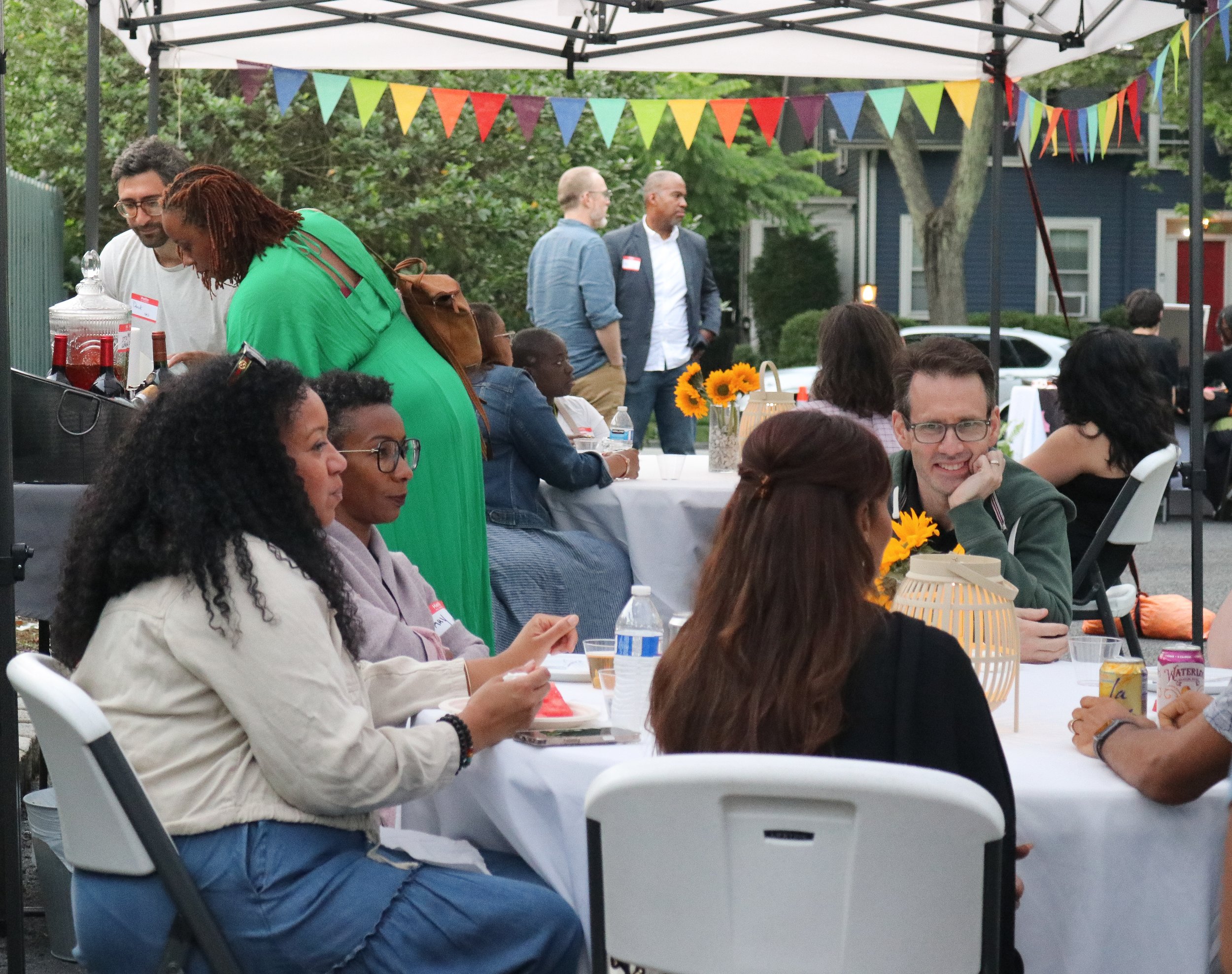 People gathered at an outdoor celebration or party decorated with colorful bunting and sunflowers, seated at tables with drinks and snacks, socializing under a canopy.