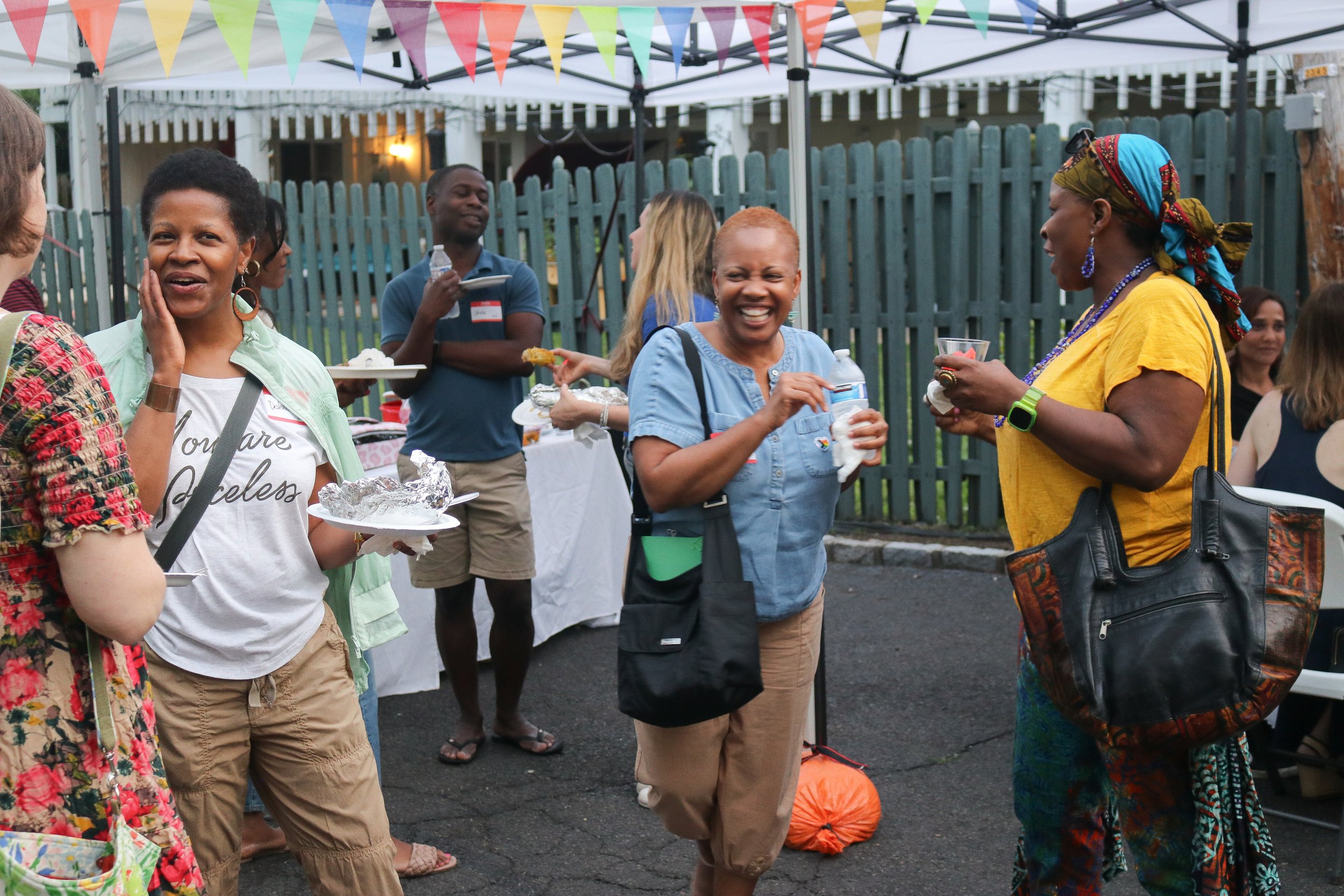 Group of diverse women and a man at an outdoor gathering under colorful banners, chatting and enjoying food and drinks.