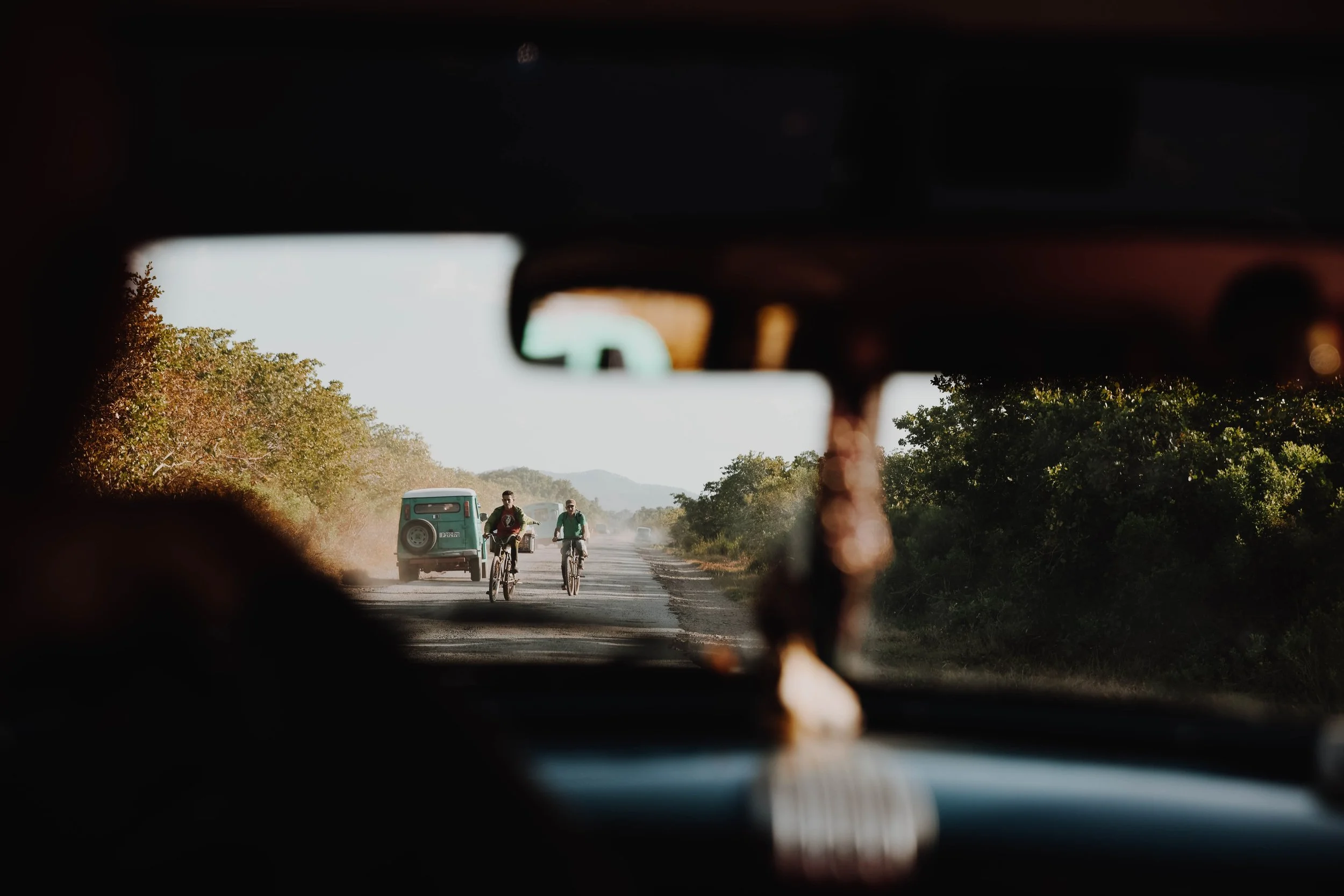 View through a car's windshield showing two cyclists riding on a dirt road, with a green auto rickshaw and another vehicle in the background, surrounded by green trees and distant hills.