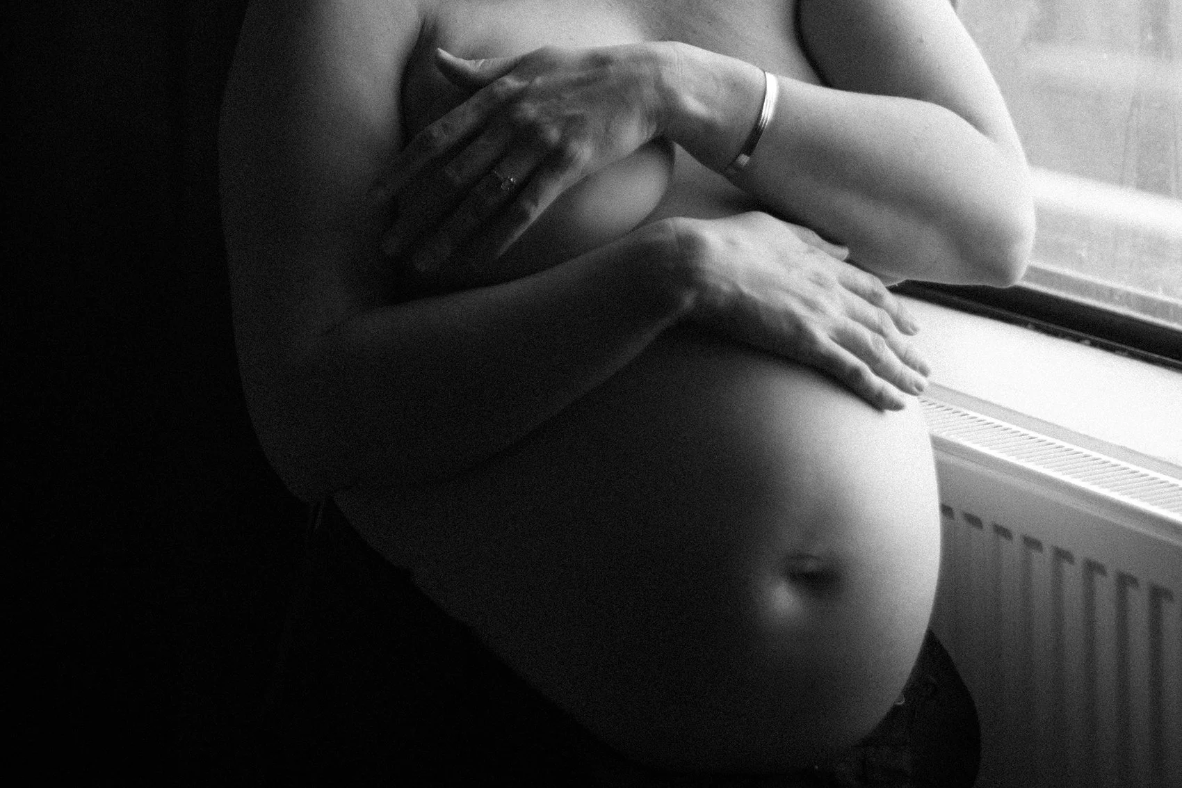 Pregnant woman sitting cross-legged on wooden floor with sheer fabric draped around her, touching her belly, in a minimalist room with textured wall.