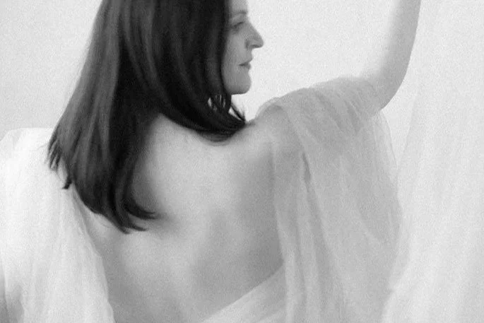 Woman with shoulder-length hair, seen from behind, in a hospital gown, sitting and leaning against a white wall or bed.