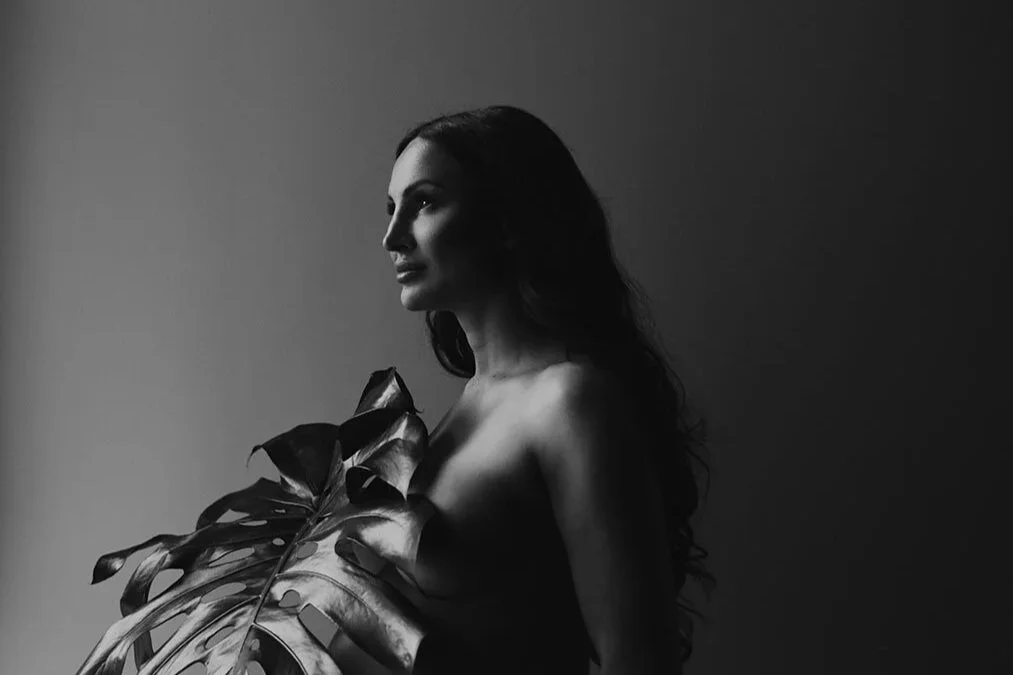 Black and white portrait of a woman with long hair holding a large tropical leaf, contrasting light and shadow.