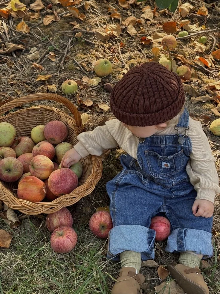 A young child sitting on the ground outdoors among fallen autumn leaves, wearing a brown knit hat, beige shirt, oversized blue denim overalls, and tan shoes. They are near a basket filled with red and green apples, with some scattered around on the g