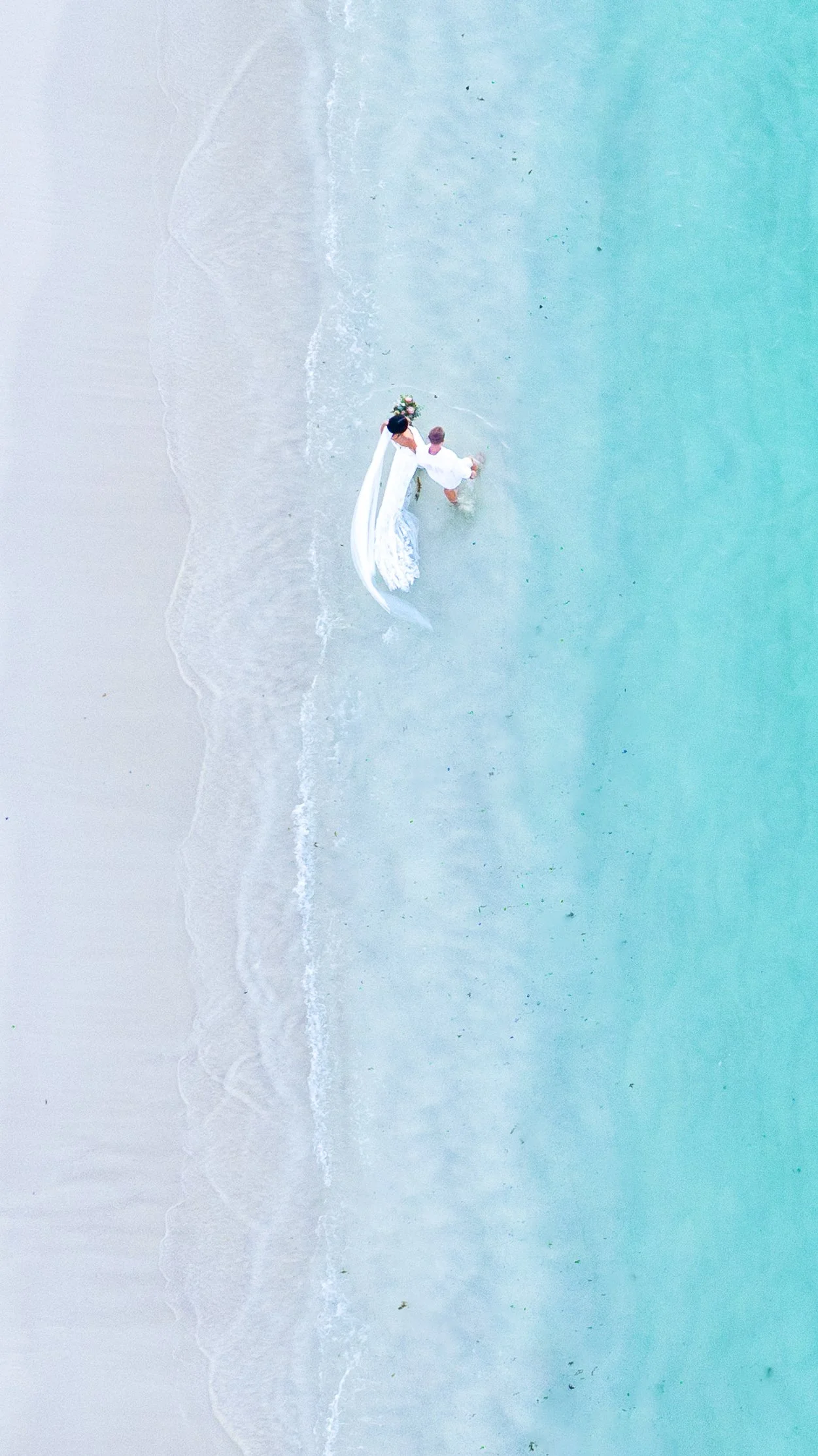 An aerial view of a couple in wedding attire standing in shallow ningaloo reef ocean water near a sandy beach, with gentle waves and turquoise water at turquoise bay