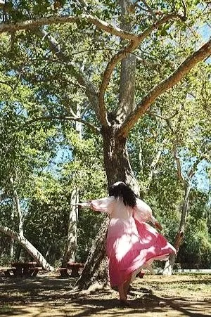 Photo of a person running away from the camera toward a tree, wearing a long pink flowing ombre dress with puffy sleeves, and long dark hair under a. head scarf.