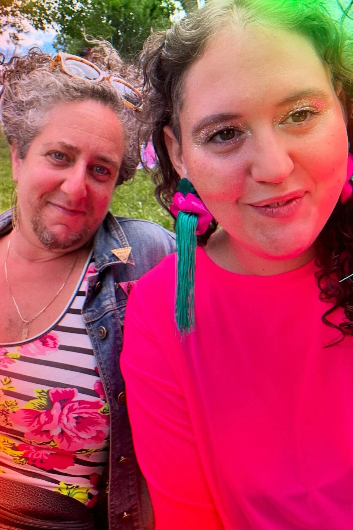 A brightly-colored slightly psychedelic photo of two light skinned curly-haired people. Their outfits include glitter, stripes, roses, hot pink and denim, lips spitting turquoise tassels, and a green flare of light in the upper right.