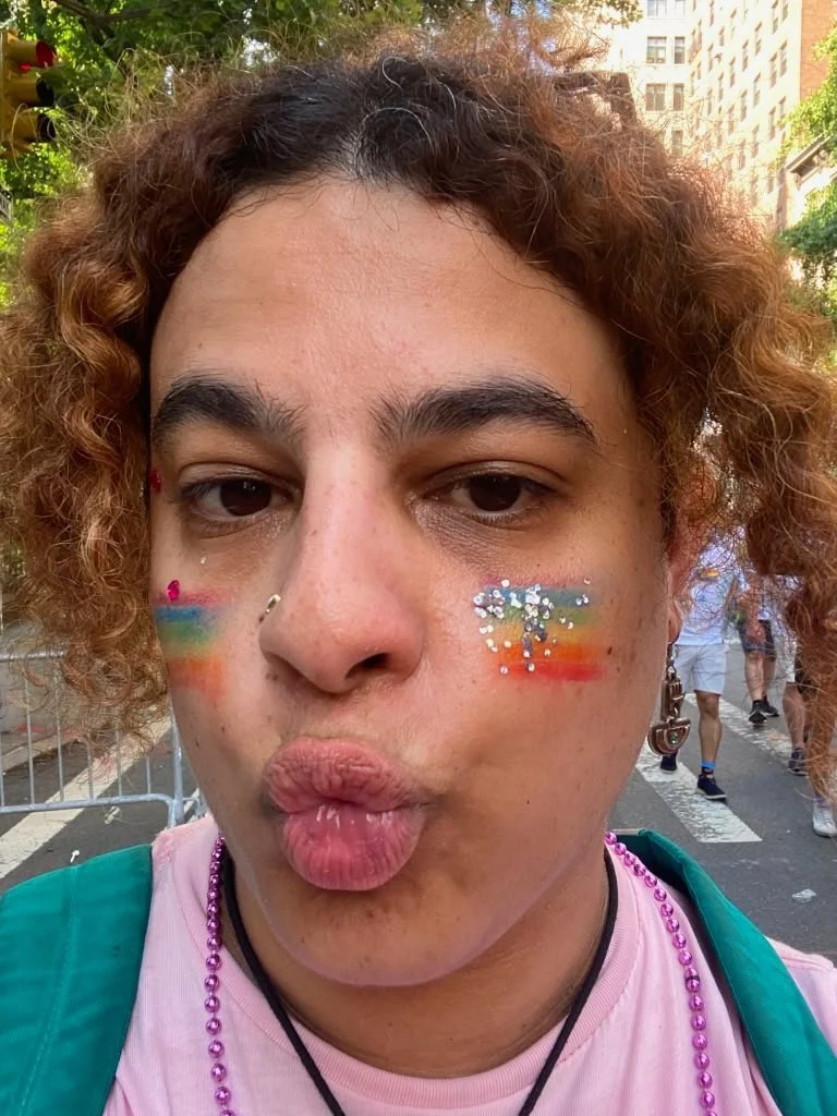 Photo of Je'Jae making a kissy face with rainbows and glitter on their face. Their hair is curly and framing their face, and are wearing a pink tshirt and purple beads.