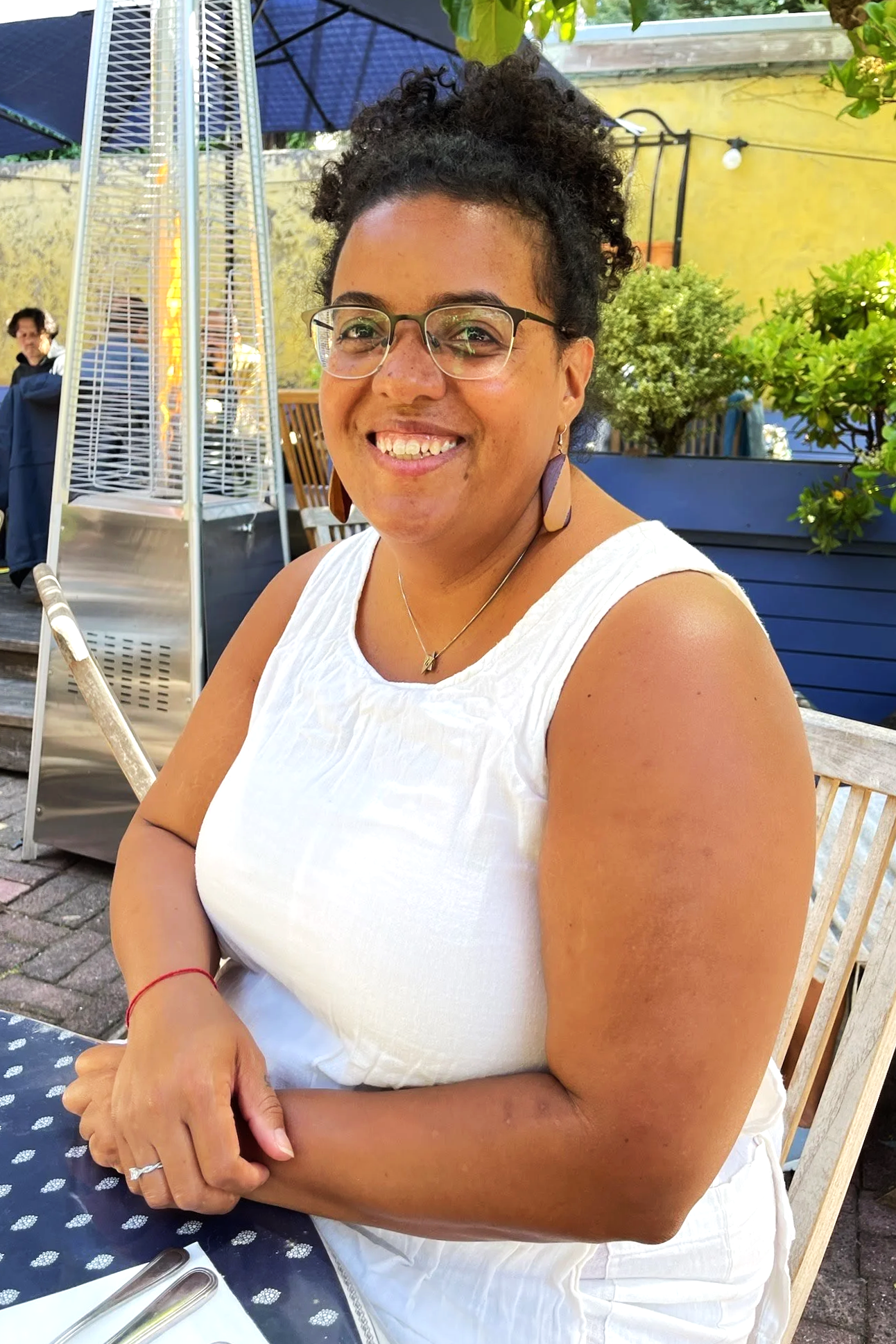 Photo of a smiling person with glasses, brown skin, a white dress, and curly hair in a ponytail on top of their head, sitting outdoors at a restaurant.
