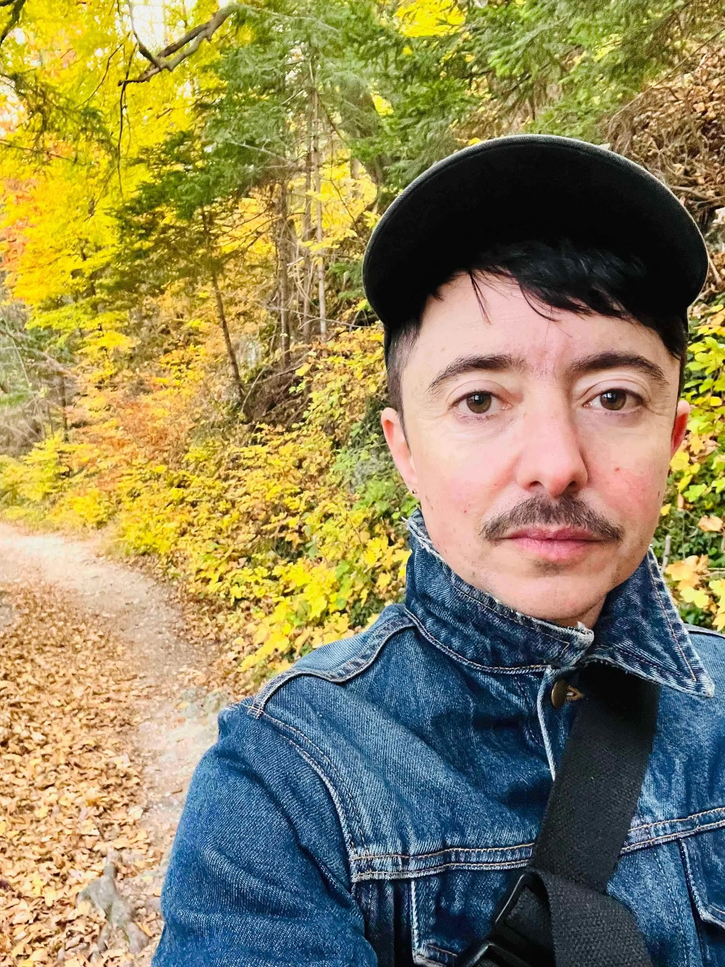 Photo of Dean wearing a hat and a jean jacket. They have a mustache, dark hair and eyes, and are on a path in the woods with yellow green foliage.