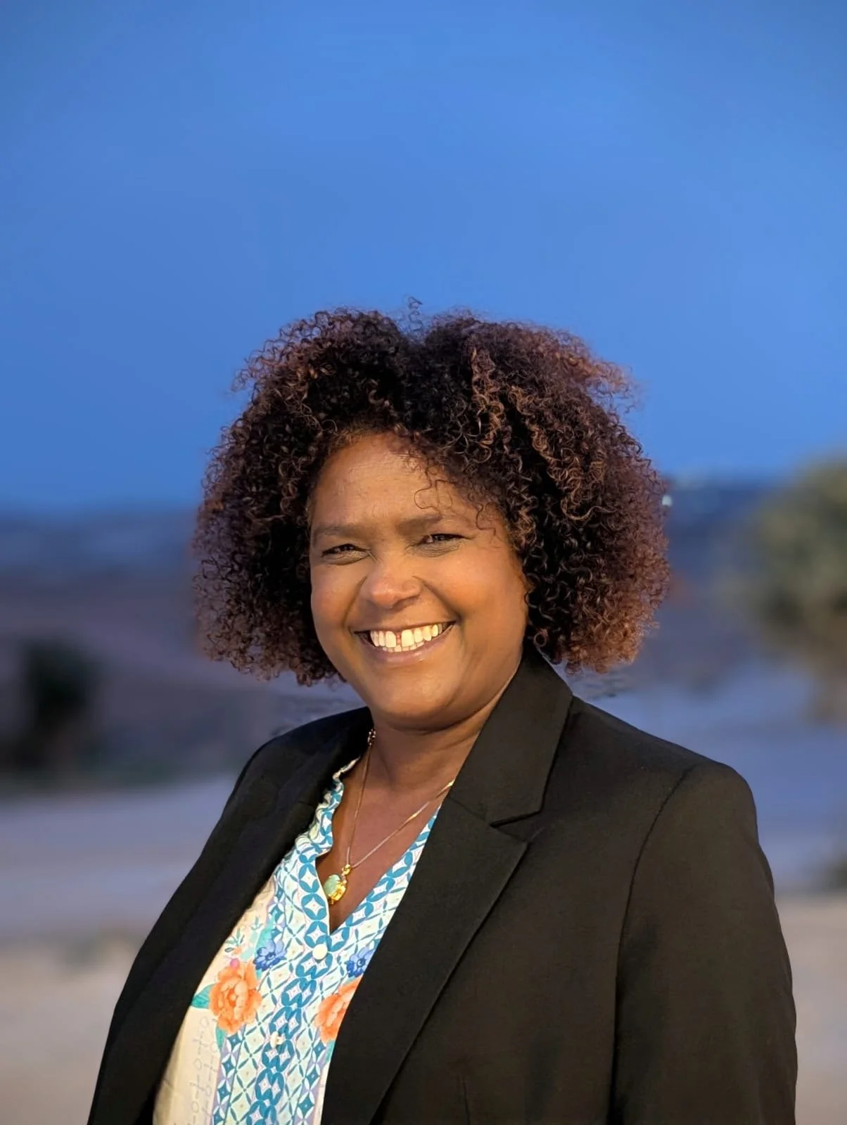 Photo of Shula wearing a black blazer over a shirt with floral and geometric designs in blue and orange. Her hair is natural, bouncy and chin length. She is smiling in front of a landscape with a bright blue sky.