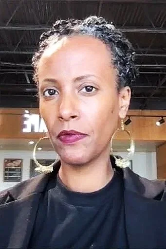 Photo of a person with brown skin, magenta lipstick, a black shirt and blazer, and big gold hoops. They are not smiling. Their hair is natural, pulled back, with salt & pepper graying.