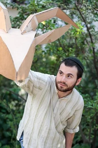 Photo of a person with light skin, dark hair, a mustache and short beard, wearing a white shirt with vertical stripes. They are holding a goat head made out of cardboard and looking at it like it has its own life.