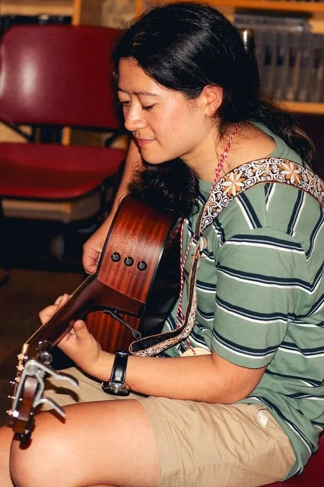 Photo of a person playing the guitar. They have long black hair pulled back in a ponytail, light brown skin, and are wearing a striped t-shirt, shorts, and a patterned guitar strap.