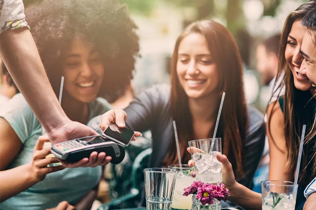 Group of four women sitting outdoors at a table, smiling, with drinks and straws, as a person accepts a credit card and device for payment.