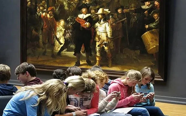Group of young girls sitting on the floor in front of a large painting in an art museum, looking at their phones.