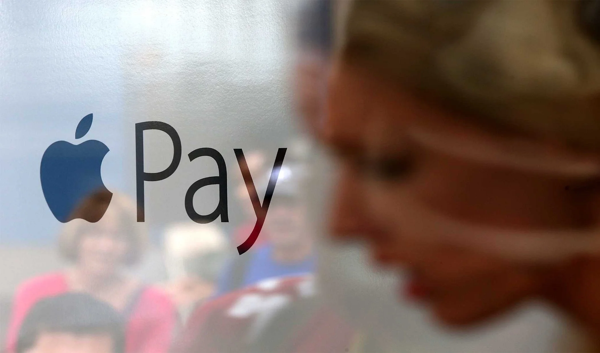 Close-up of an Apple Pay logo on a glass surface with blurred people in the background.