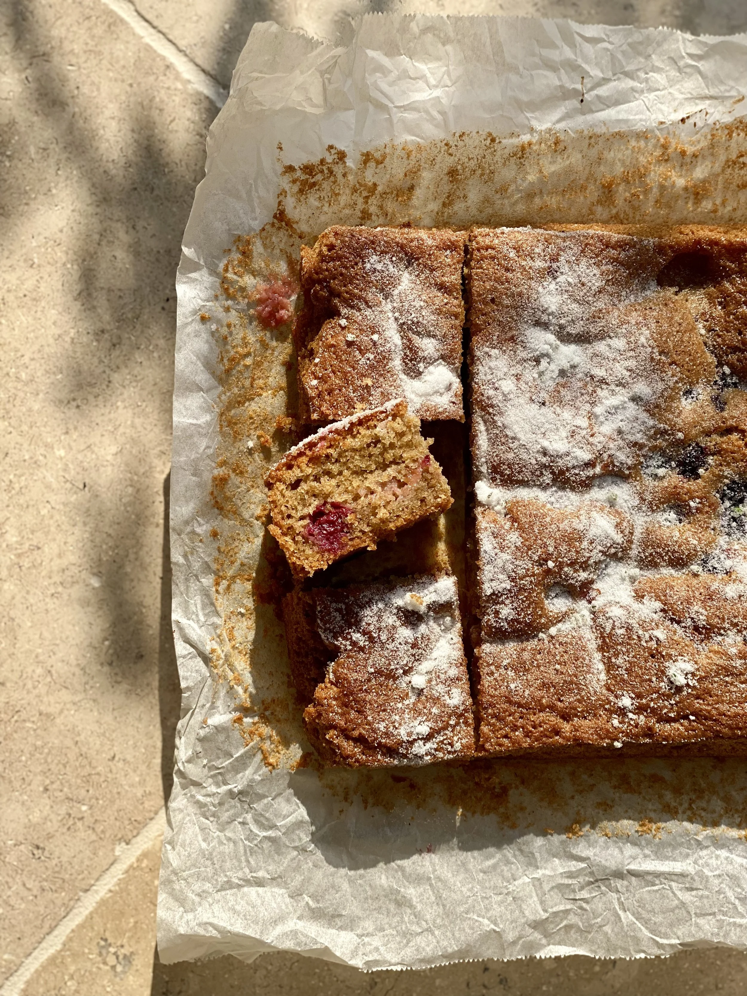 A freshly baked blackberry and plum cake dusted with icing sugar on parchment paper outdoors.