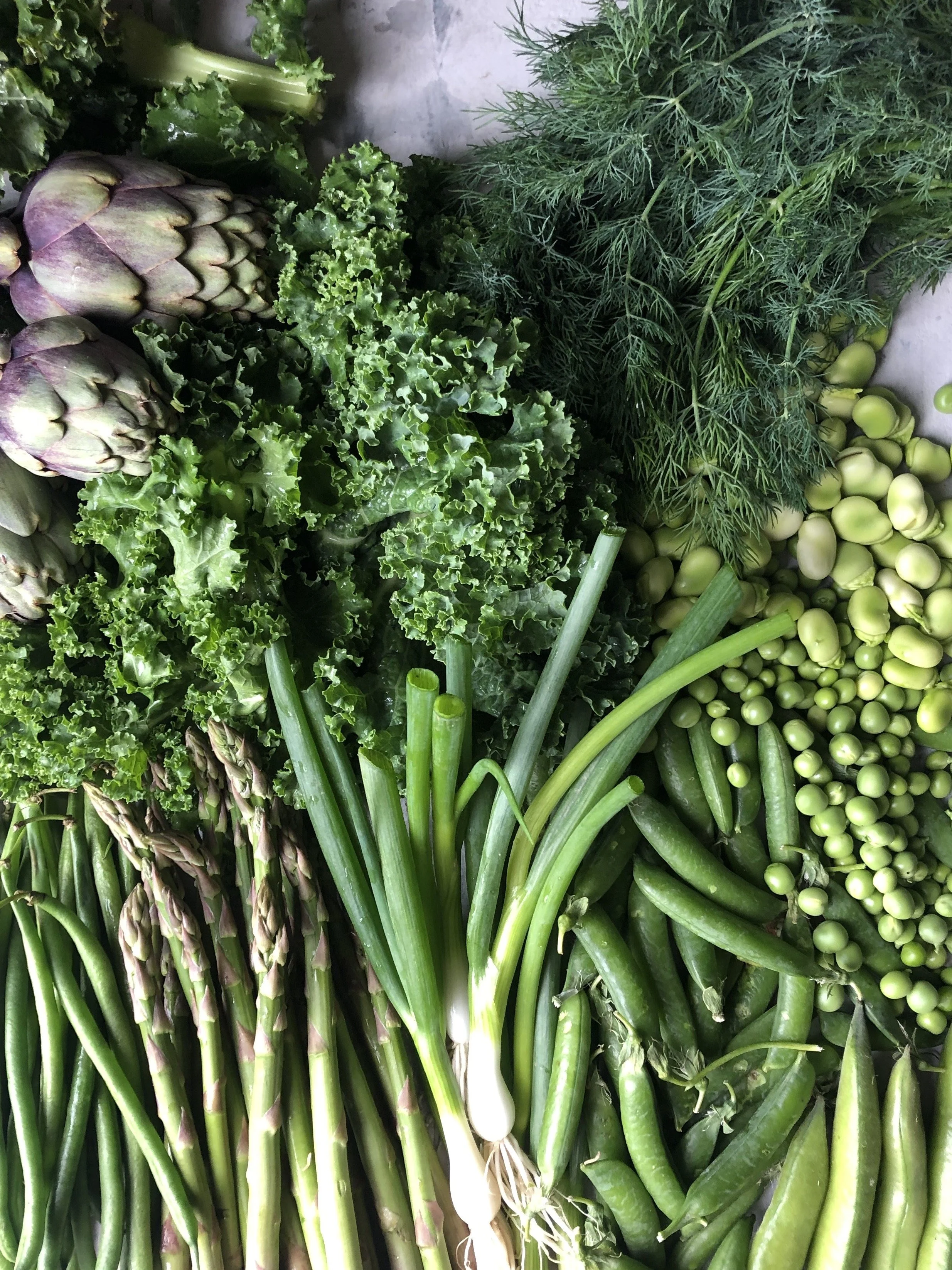 Fresh green vegetables including artichokes, kale, dill, tomatoes, green beans, asparagus, and cucumbers.