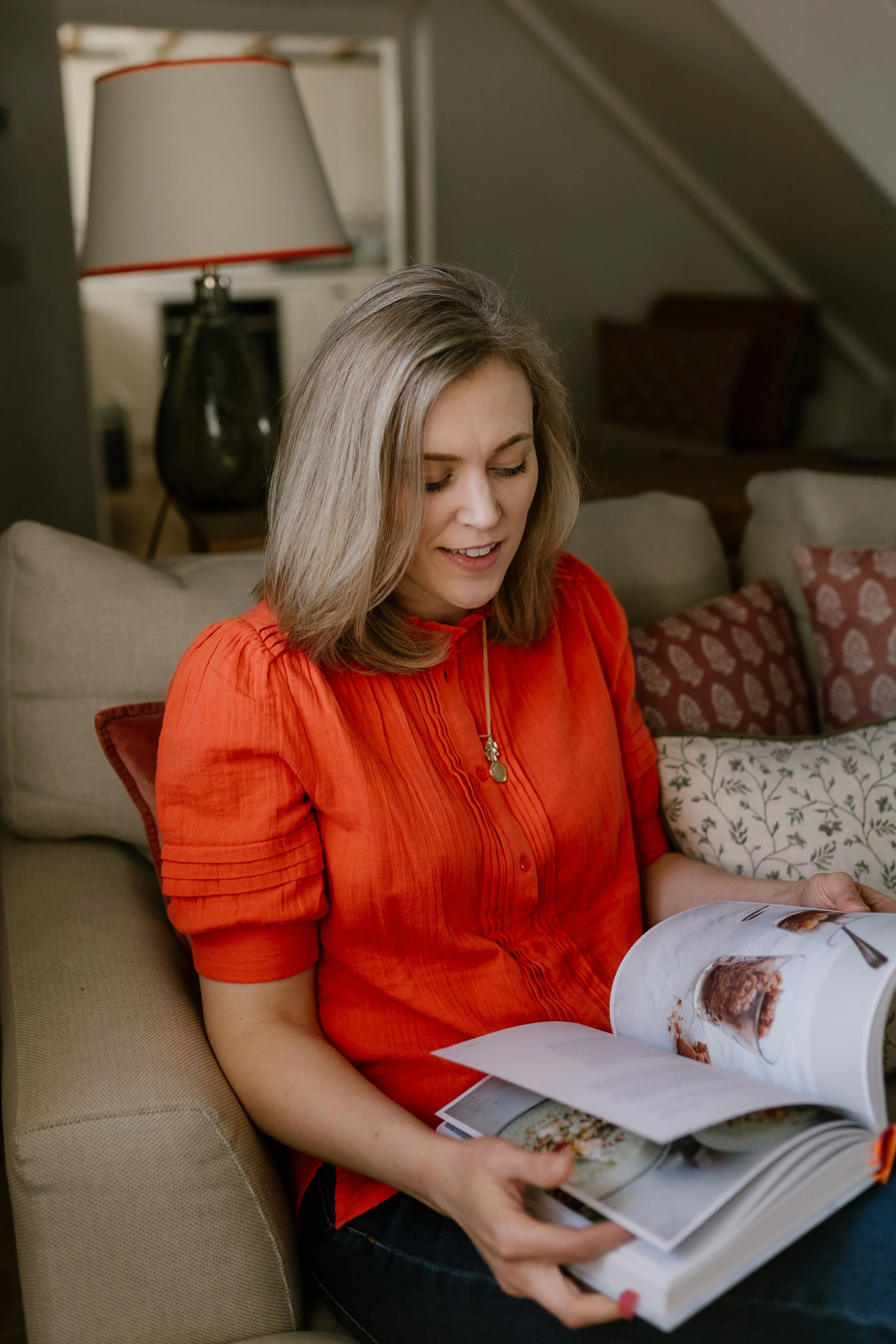 A woman with blonde hair, wearing an orange top, is sitting on a beige couch and looking at a large photo book with a meal or dessert picture.