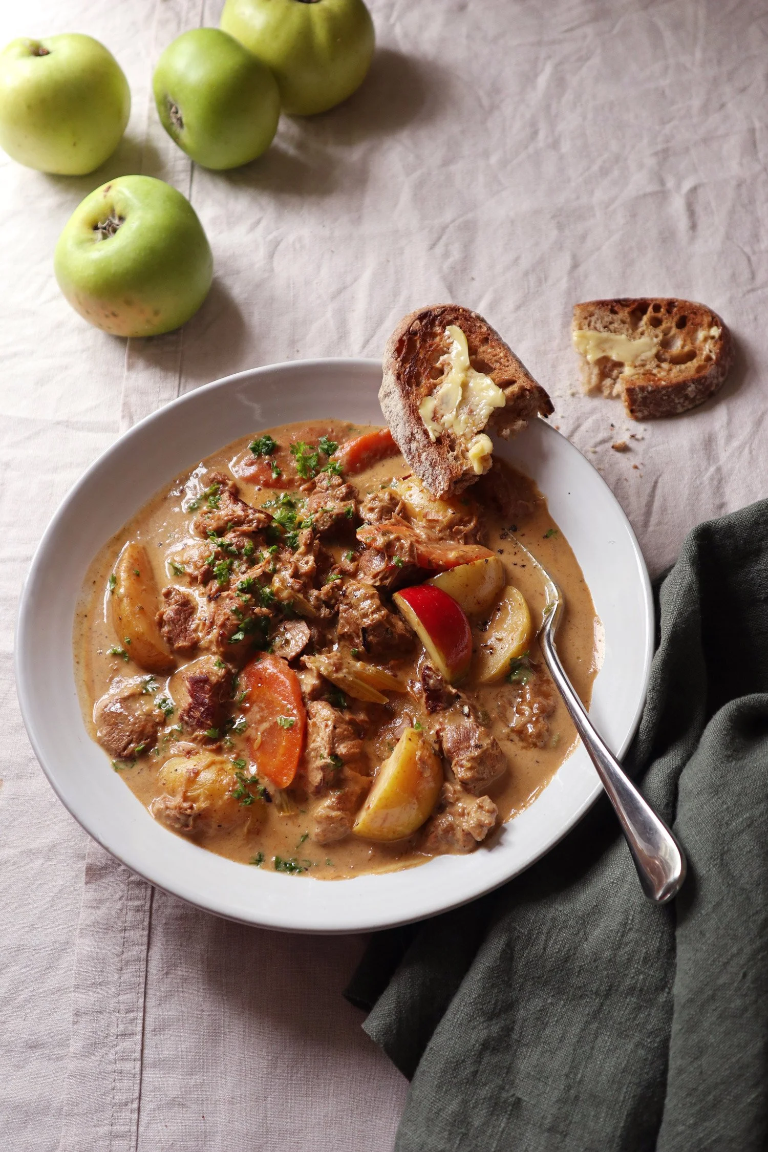 A bowl of pork stew with carrots, potatoes, and herbs, served with slices of bread with butter, on a table with green apples and a black napkin.