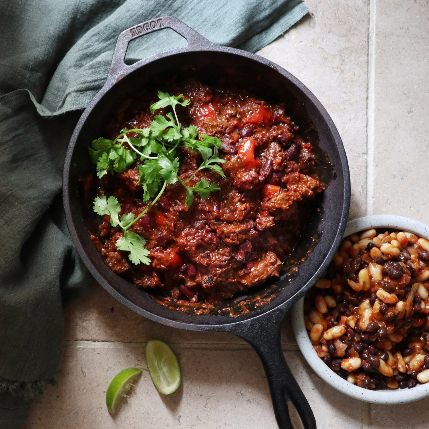 A cast iron skillet filled with hearty chilli topped with fresh coriander, next to a bowl of baked beans and a lime wedge on a light-coloured surface.