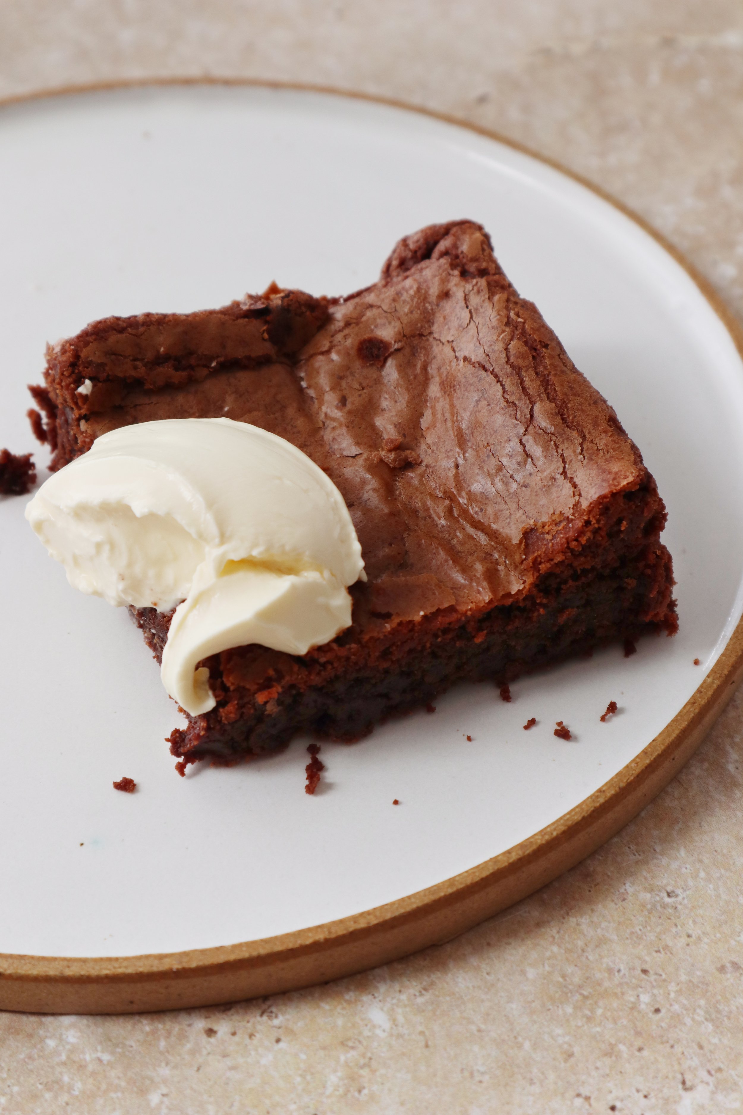 A slice of chocolate brownie with a dollop of vanilla ice cream on top, served on a white plate with a wooden rim.