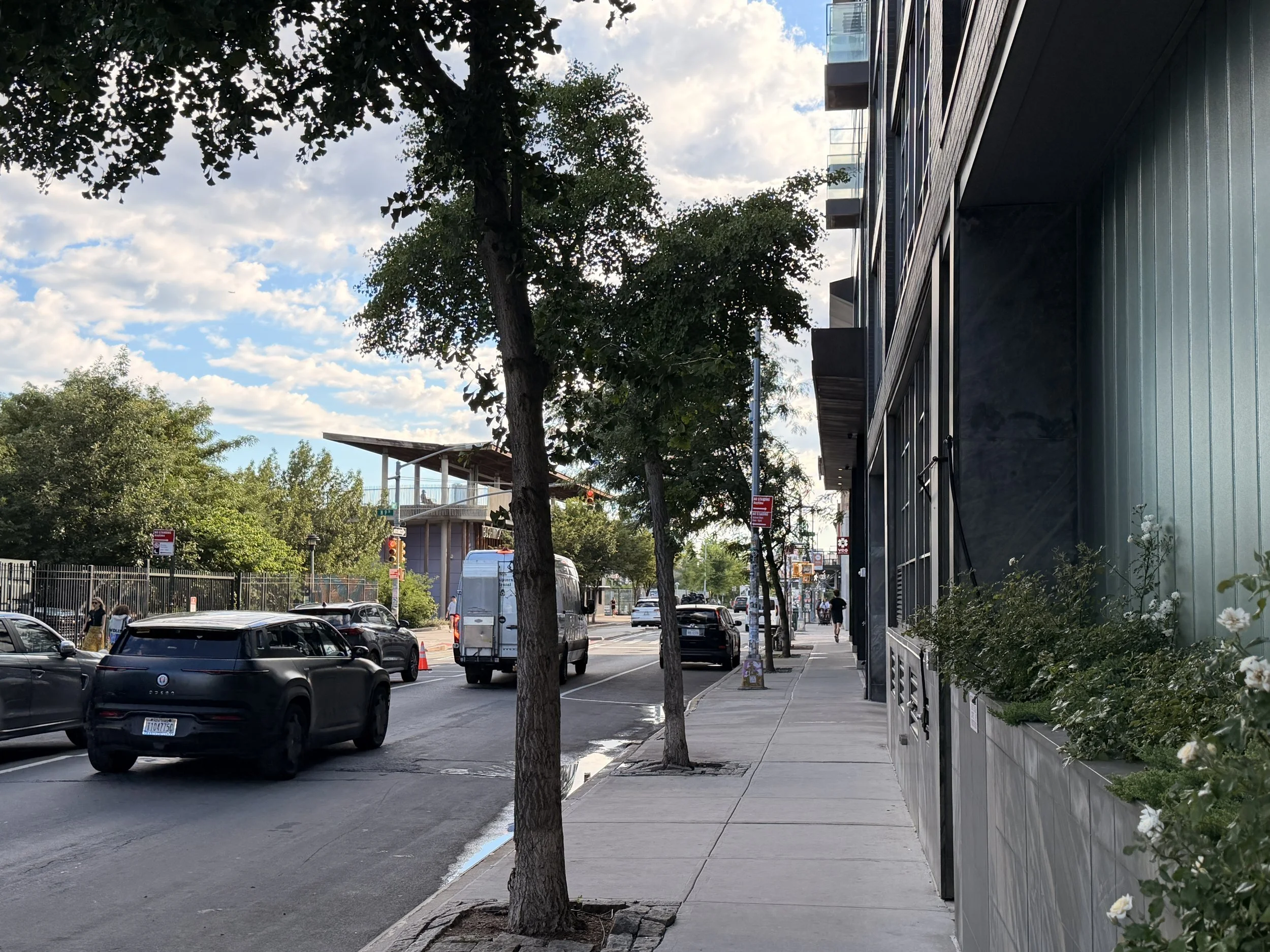 City street scene with parked cars along the curb, trees lining the sidewalk, and modern buildings with glass balconies on the right. The sky is partly cloudy with patches of blue, and there are pedestrians walking on the sidewalk.