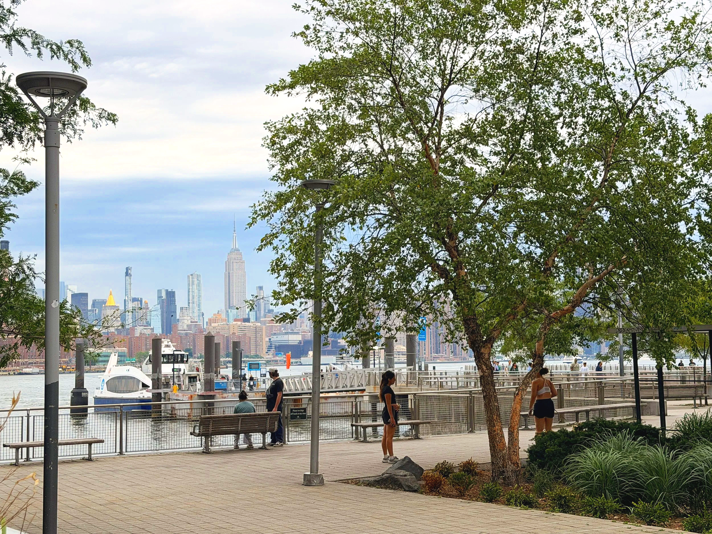City skyline visible over waterfront park, with trees and benches in foreground, and people walking along the waterfront.