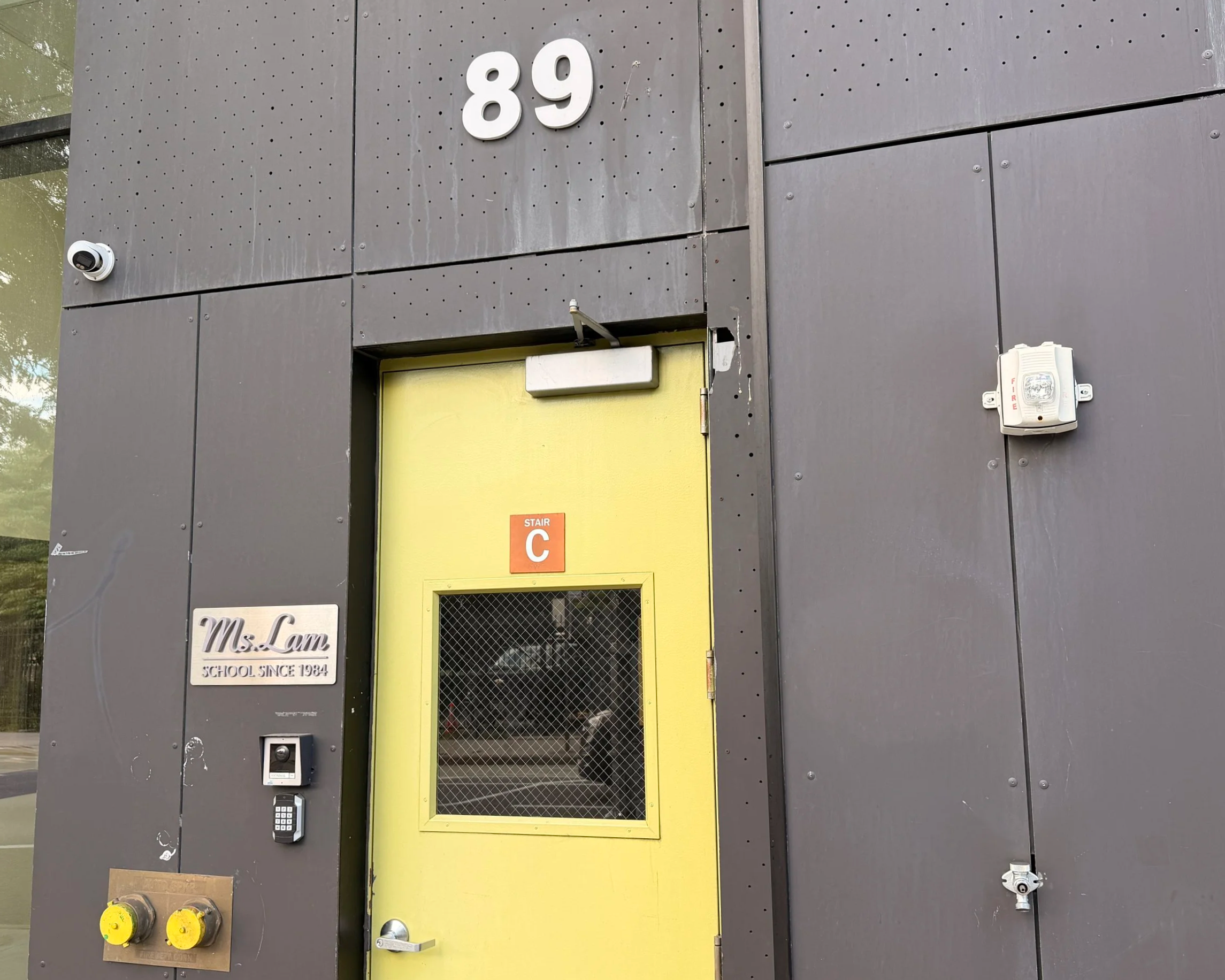 A building entry door with the number 89 displayed above it. The door has a yellow frame and a peep hole, with a sign that says 'Ms. Lam School Since 1934'. There are security devices including an intercom, a keypad, a surveillance camera, and a motion sensor on the wall near the door.