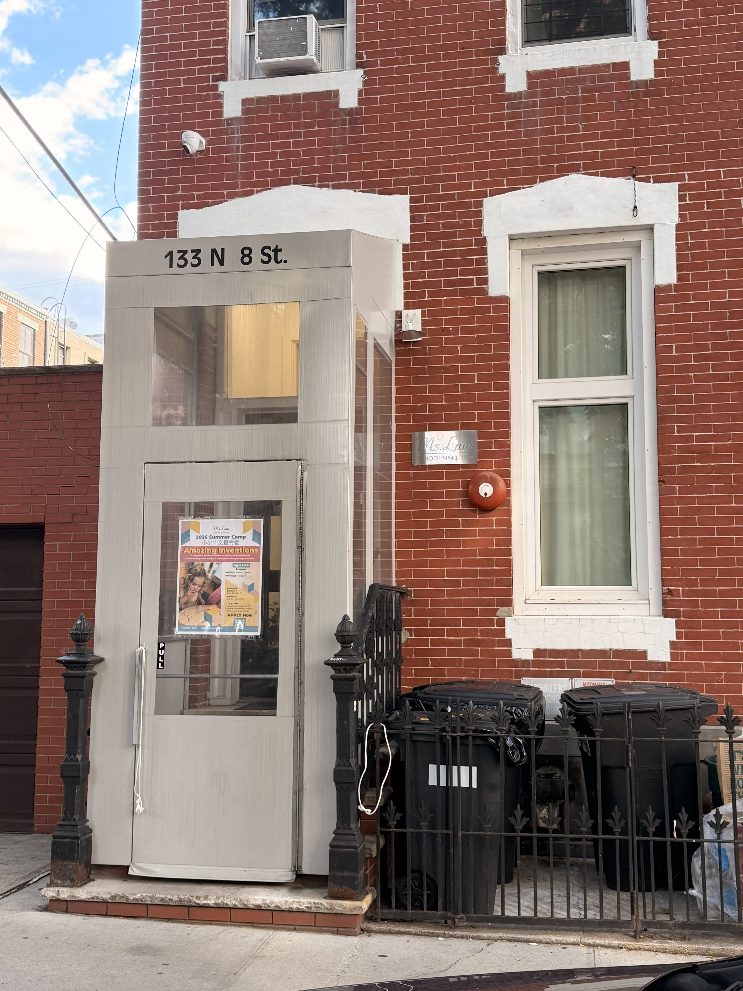 A red brick building with white trim around windows and a white entrance structure displaying the address 133 N 8 St. There are three black trash bins and a black metal fence in front of the building. A poster is on the door, and a small silver sign reads 'Ms. Lam', indicating a school. The building has an air conditioning unit and a security camera installed.