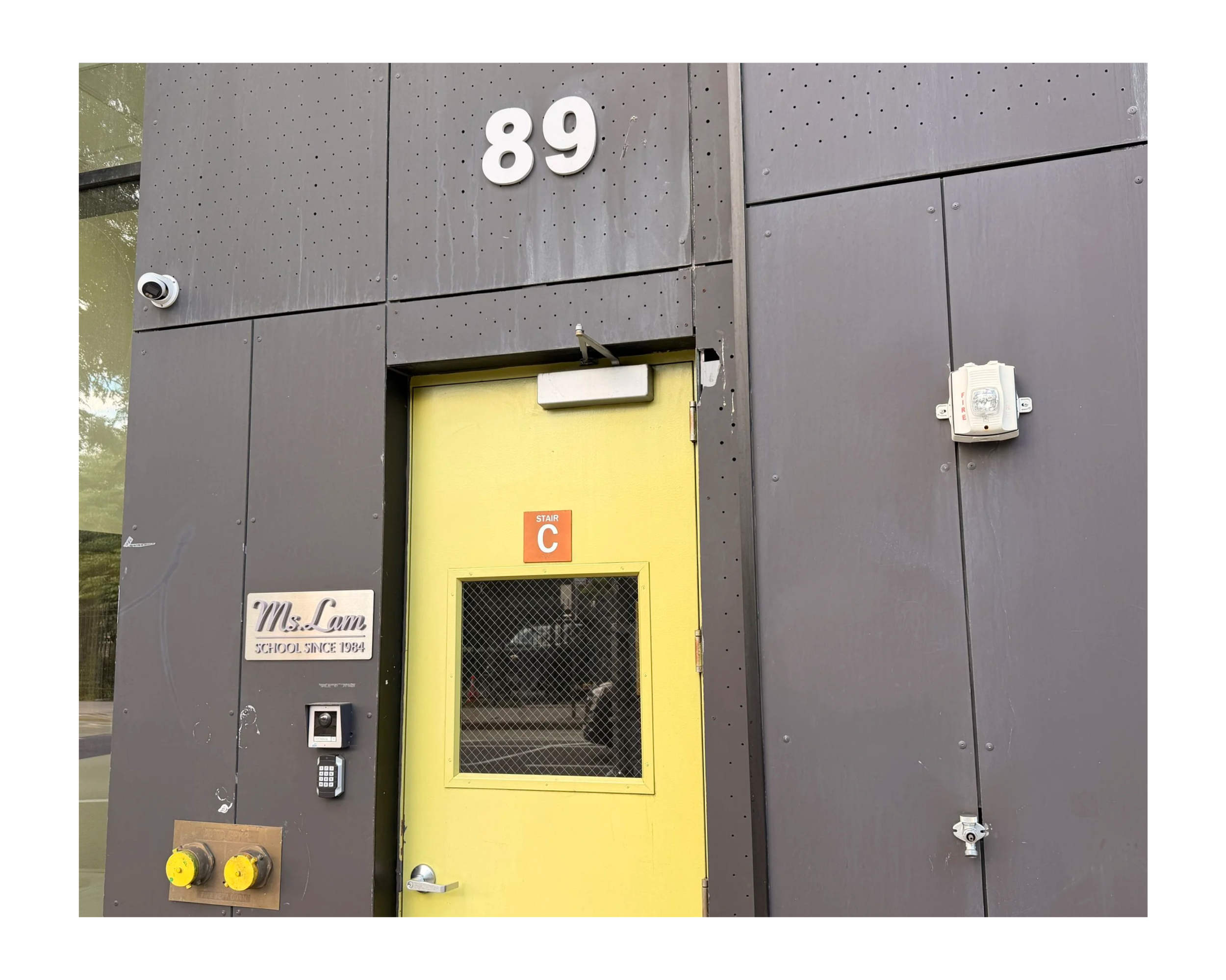A modern building exterior with a gray metal wall, a yellow door with a wire mesh window, and the number 89 on the wall. There is a security camera, an intercom, and a fire alarm box on the wall, along with a small sign reading "Ms. Lam School since 1984."