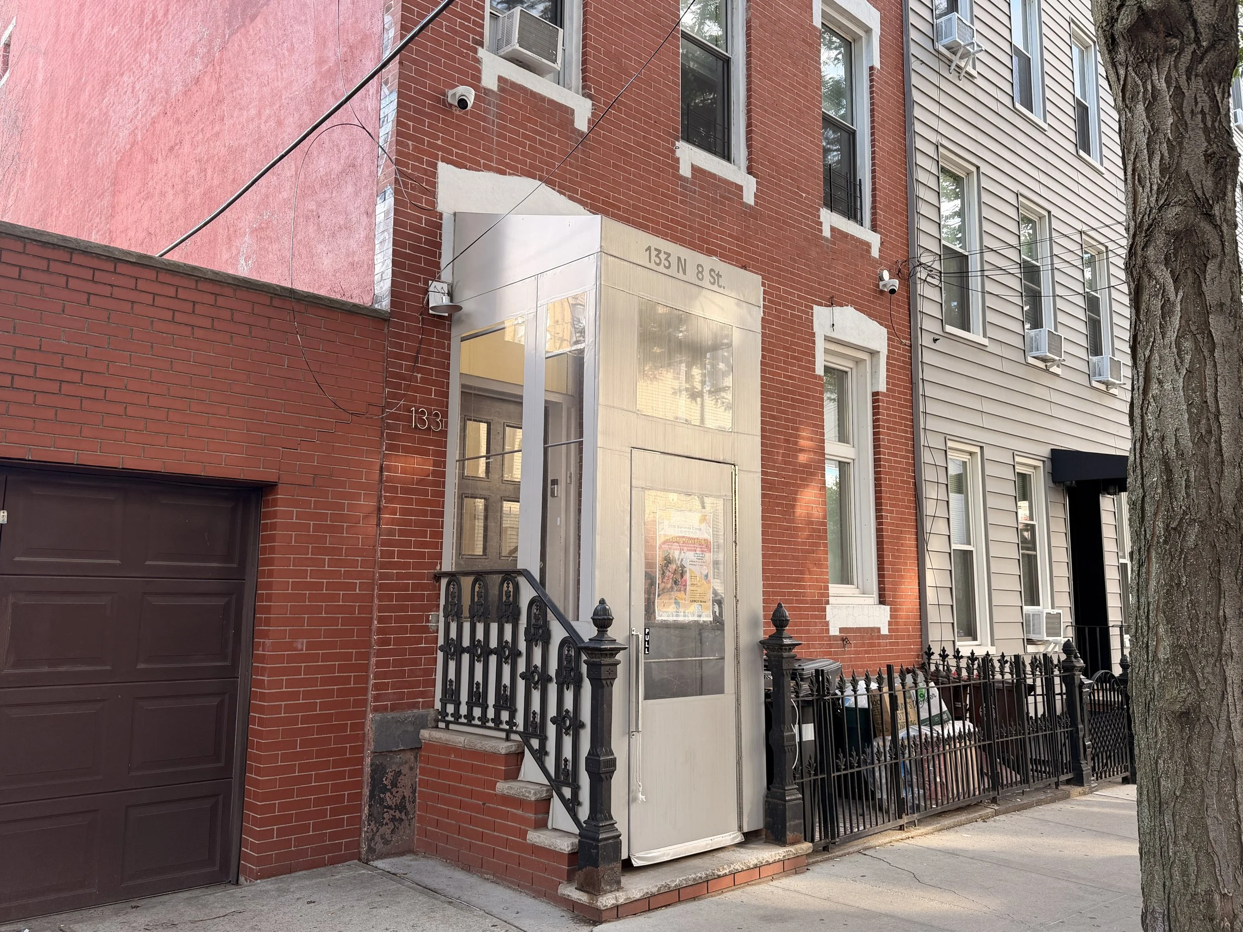 Street view of a row of brick and siding houses with black iron fences, a tree, and a sidewalk.