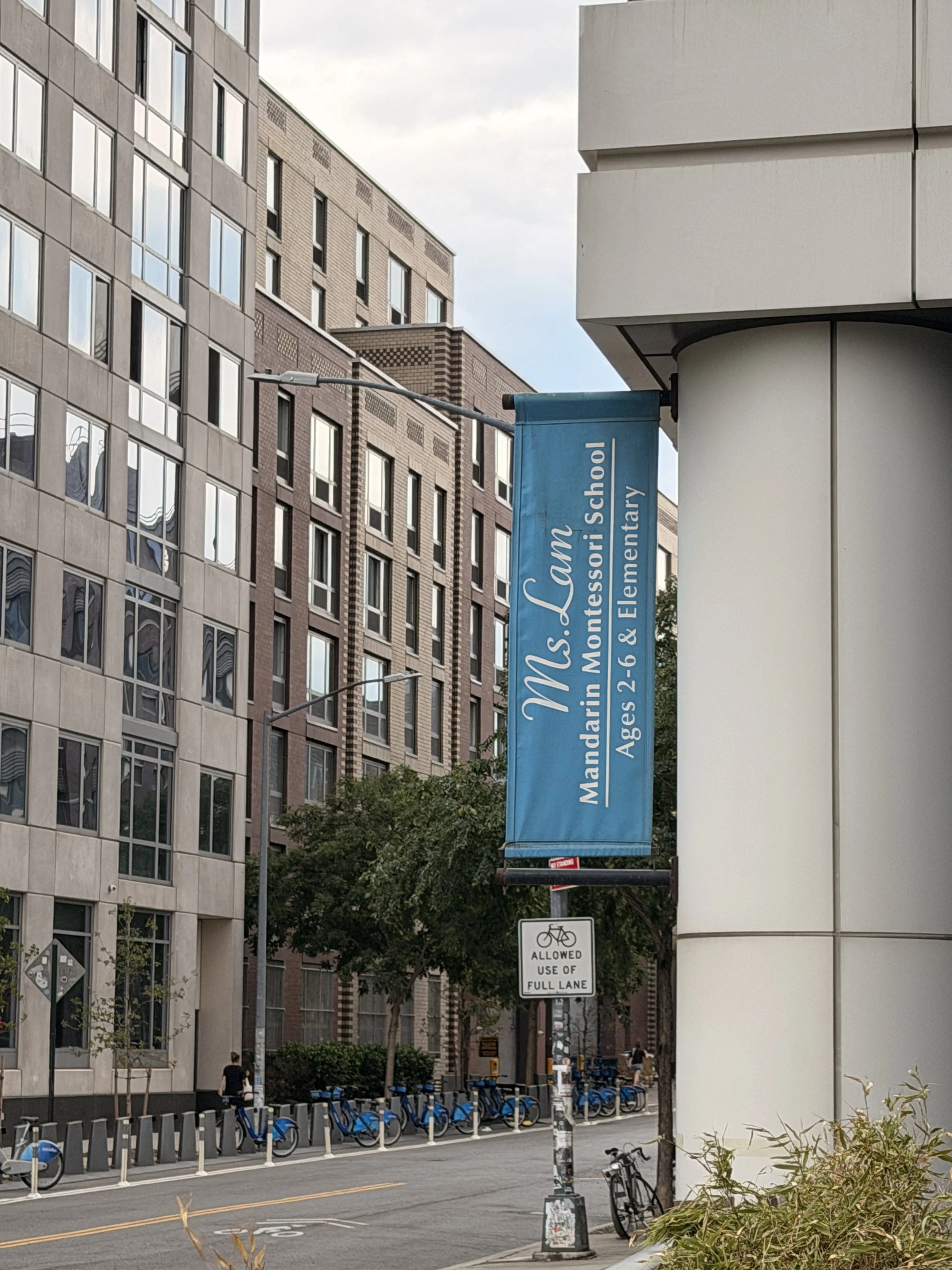 Street scene with modern buildings, a row of blue bike-sharing bicycles, a sign indicating bicycle use is allowed in a full lane, and a blue banner for Mandarim Ms. Montessorii School.
