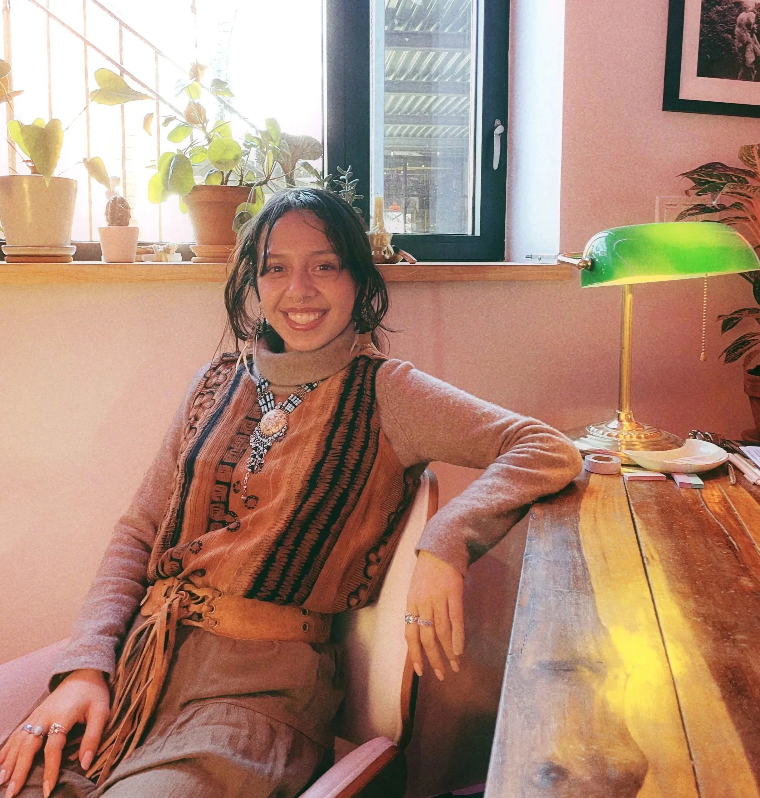 A woman with dark, curly hair smiling and sitting in a cozy room, wearing layered necklaces and a patterned vest over a tan turtleneck, with a window and several potted plants behind her, and a wooden desk with a green-shaded lamp and various objects on it.
