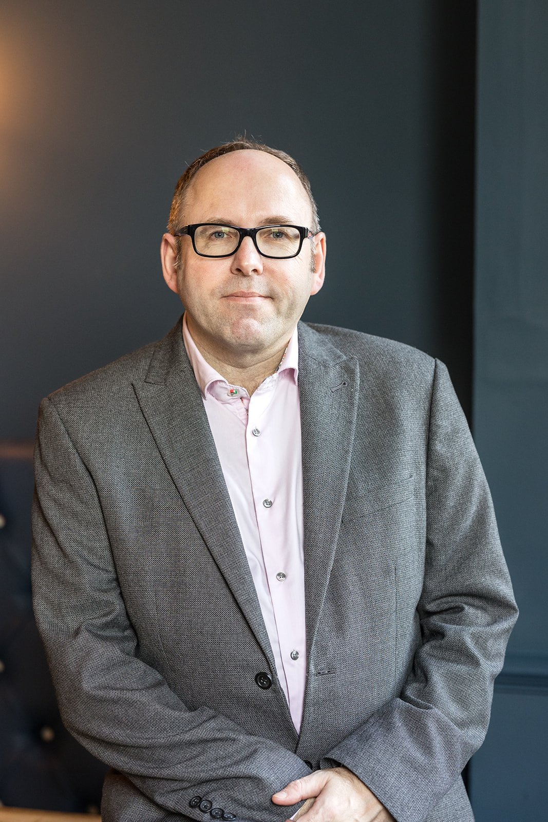 A professionally dressed man with glasses, dressed in a jacket and open necked shirt.