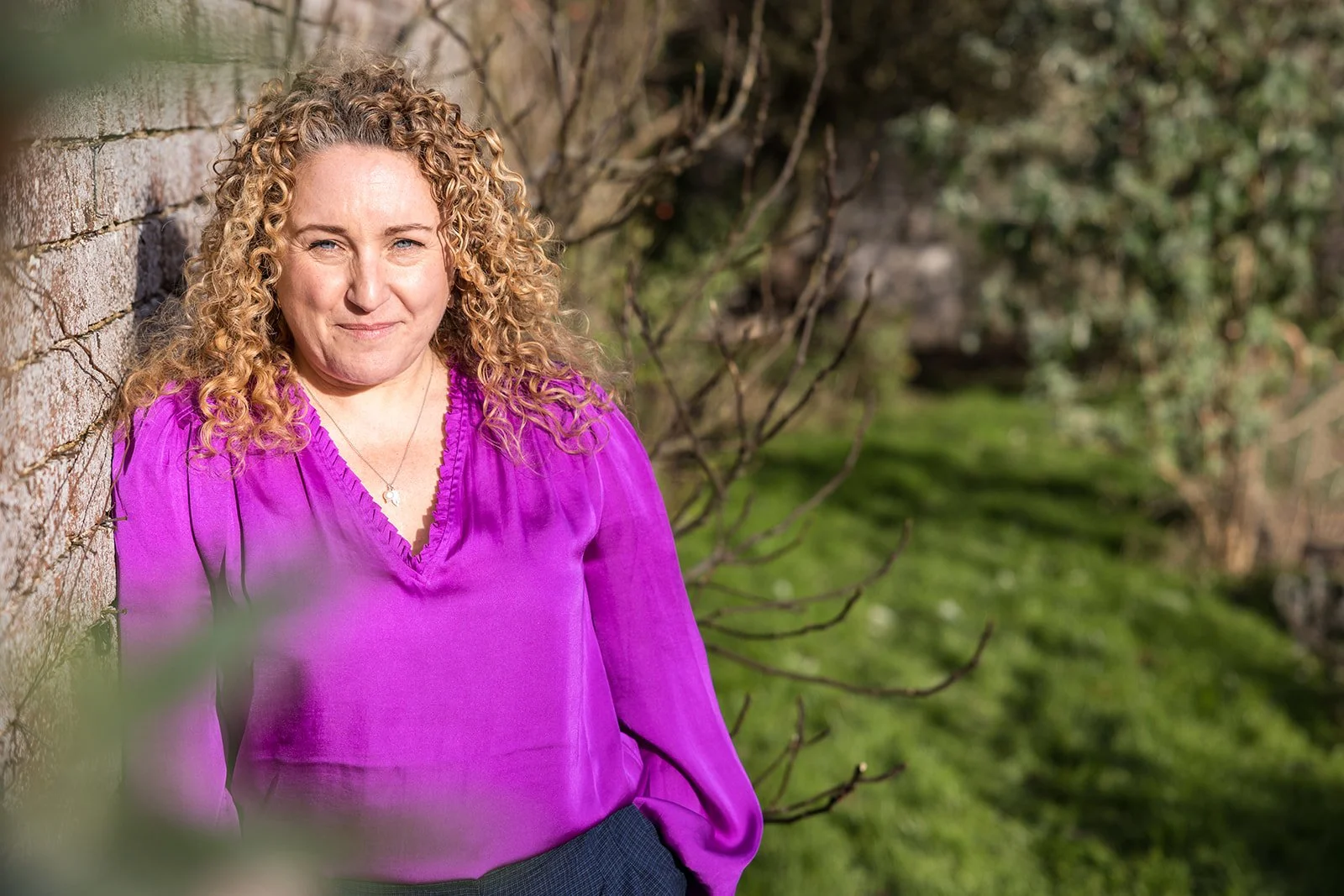 A woman with curly blonde hair leaning against a brick wall outdoors, wearing a purple blouse and smiling.