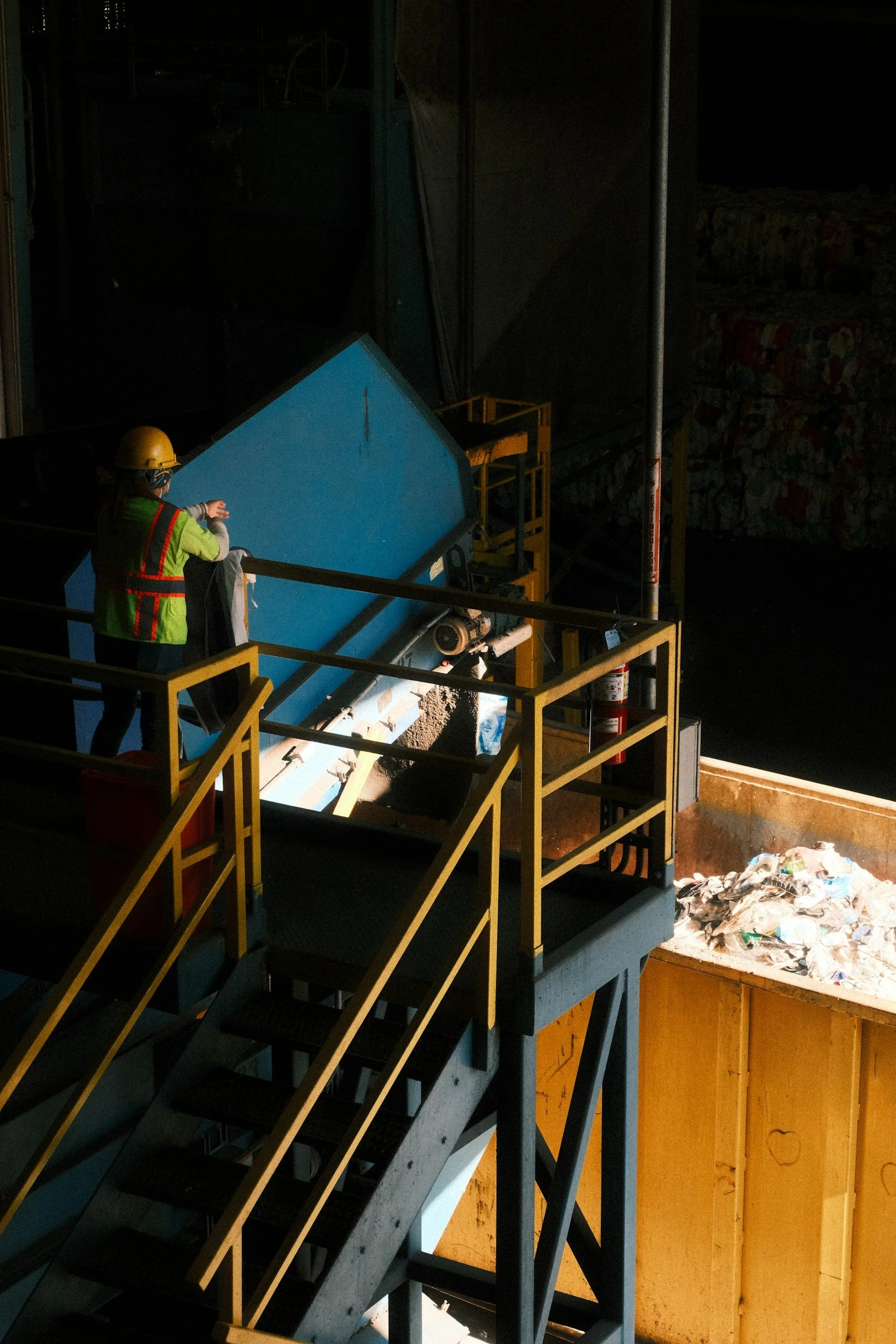 A man dressed in PPE and standing on steps, looking down on a skip of waste paper.