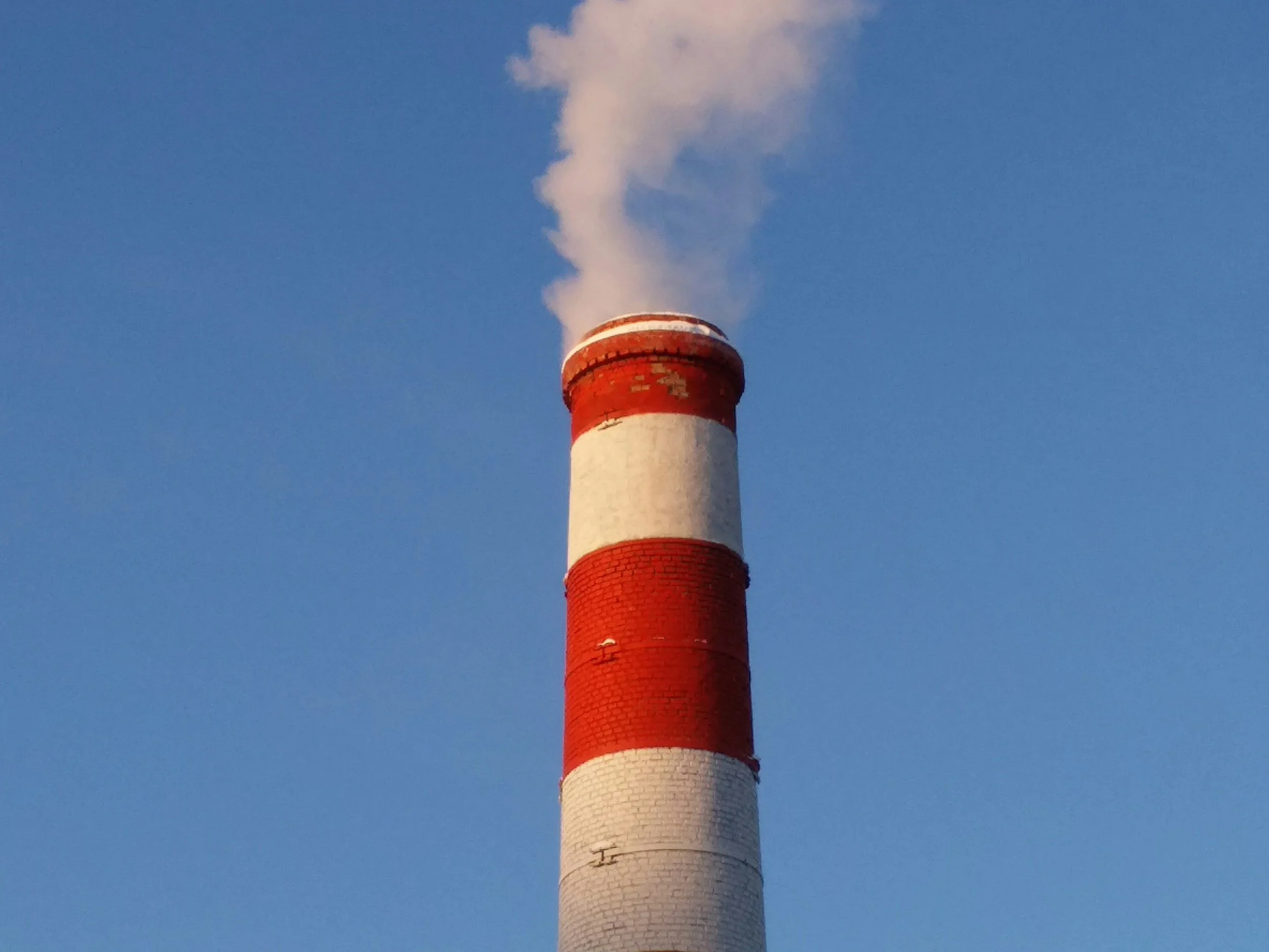 A tall industrial chimney with red and white stripes emitting white smoke into a clear blue sky.